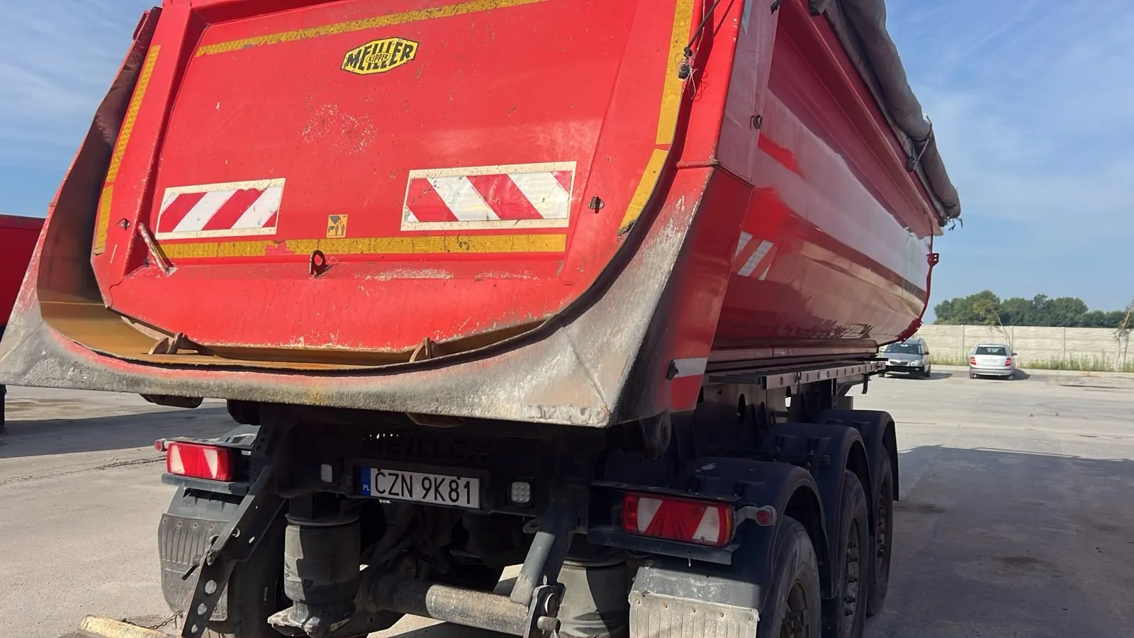 Tylny widok czerwonej naczepy wywrotki marki MEILLER. Na tylnej klapie znajdują się dwa biało-czerwone pasy ostrzegawcze oraz logo producenta. Poniżej widoczna jest tablica rejestracyjna o numerze CZN 9K81. Naczepa posiada trzy osie z kołami. W tle widać betonowy mur i dwa zaparkowane samochody osobowe na jasnym placu pod niebem bez chmur.