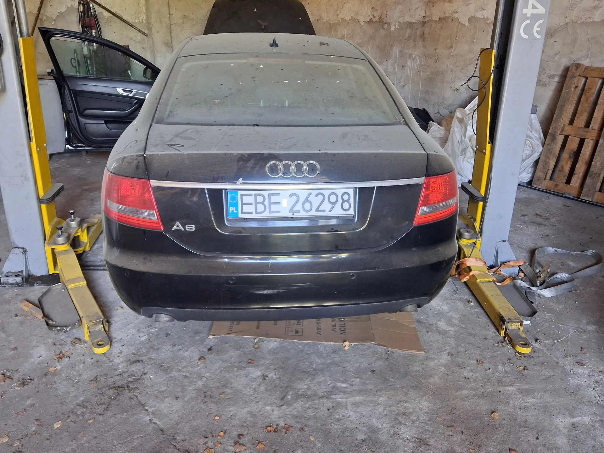 Tylny widok czarnego samochodu marki Audi A6 z polską tablicą rejestracyjną EBE 26298. Samochód jest zakurzony i znajduje się na żółtym podnośniku warsztatowym w garażu. Lewe tylne drzwi są otwarte.