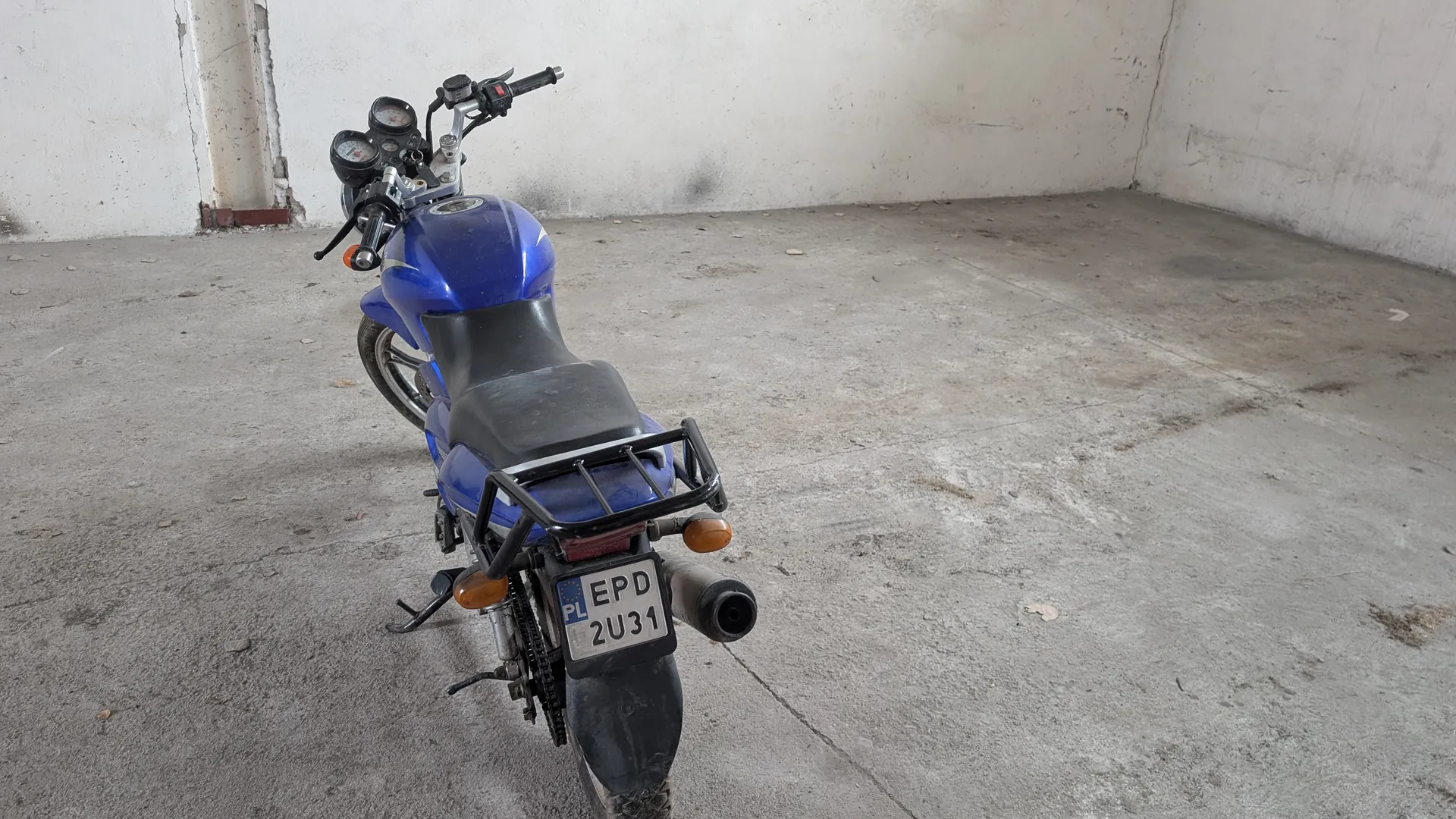 Niebieski motocykl widziany od tyłu, stojący na betonowej podłodze w pustym pomieszczeniu z białymi ścianami. Widoczna czarna siedziba, czarny bagażnik, pomarańczowe kierunkowskazy oraz tablica rejestracyjna z napisem PL EPD 2U31. Na kierownicy znajdują się wskaźniki i czarne uchwyty. Podłoga i ściany mają ślady zużycia.