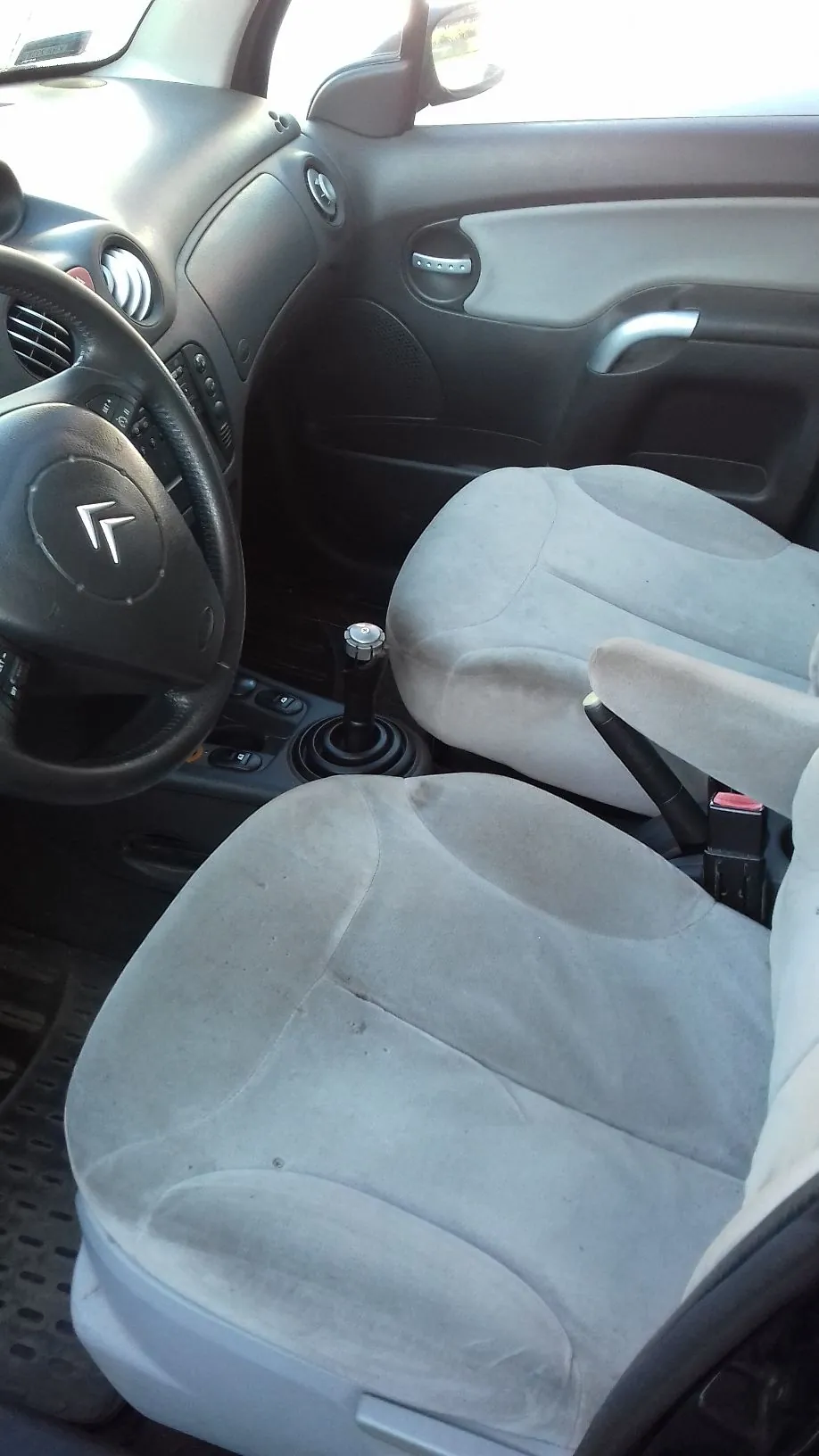 Wnętrze samochodu z perspektywy kierowcy. Widoczna jest czarna kierownica z logo Citroëna, deska rozdzielcza, lewy fotel pasażera oraz przedni fotel kierowcy, oba obite jasnoszarą tkaniną. Pomiędzy fotelami znajduje się lewarek zmiany biegów oraz hamulec ręczny.