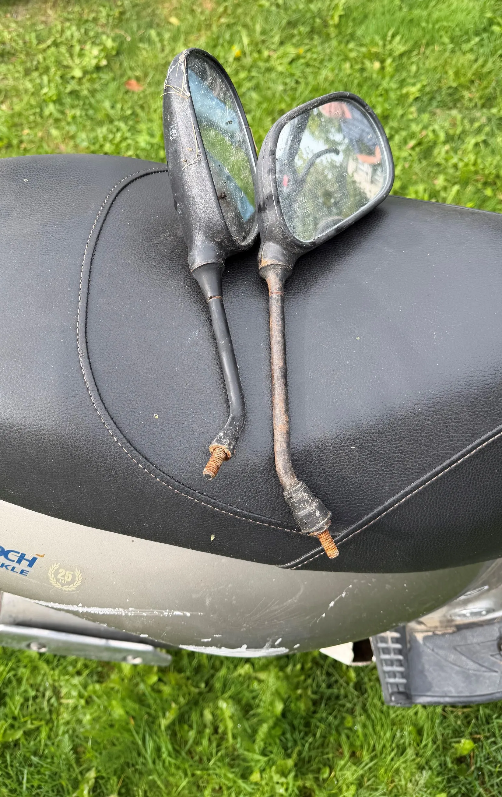 Dwa czarne lusterka motocyklowe leżące na czarnym, skórzanym siedzeniu motocykla. Lusterka mają metalowe ramiona, z których jedno jest wyraźnie zardzewiałe. W tle widoczna jest zielona trawa oraz fragment srebrnego nadwozia motocykla z widocznym fragmentem napisu 'CH KLE' oraz logo z liczbą '25'.