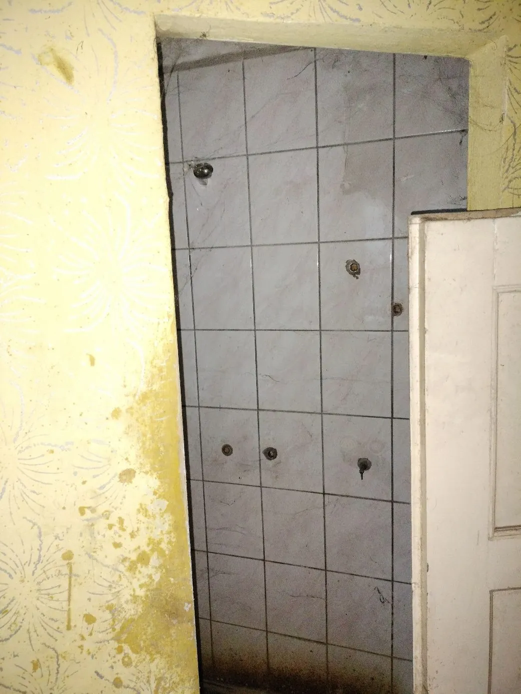 Zdjęcie fragmentu wnętrza pomieszczenia. Po lewej stronie widoczna jest żółta ściana z wytartą tapetą o kwiatowym wzorze i widocznymi zabrudzeniami. W centrum znajduje się wnęka wyłożona jasnoszarymi, kwadratowymi płytkami ceramicznymi, w których widać kilka ciemnych dziur po kołkach lub wkrętach. Po prawej stronie widoczny jest fragment białych, drewnianych drzwi.