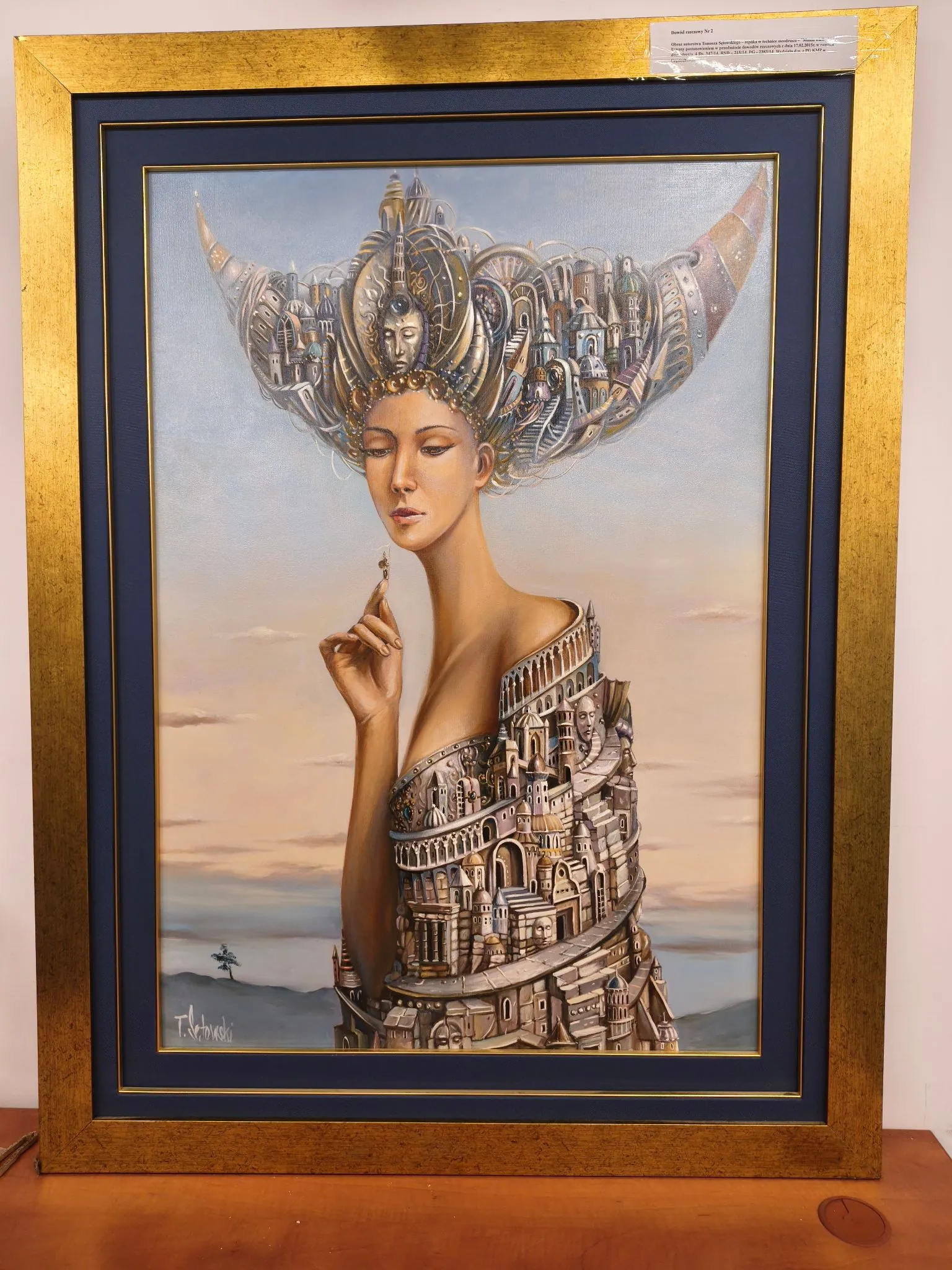 Obraz w złotej ramie z ciemnoniebieskim passe-partout, przedstawiający surrealistyczny portret kobiety. Zamiast włosów kobieta ma na głowie rozbudowaną, fantastyczną strukturę przypominającą miasto z wieżami i budynkami, uformowaną w kształt szerokiego hełmu lub korony. Jej ciało od ramion w dół również przechodzi w architekturę, tworząc wielopoziomowe miasto z łukami i schodami. Kobieta ma spokojny wyraz twarzy i uniesioną dłoń z palcem wskazującym skierowanym w górę. Tło stanowi jasne, pastelowe niebo z delikatnymi chmurami i zarysem horyzontu z pojedynczym drzewem po lewej stronie. W lewym dolnym rogu znajduje się podpis autora.