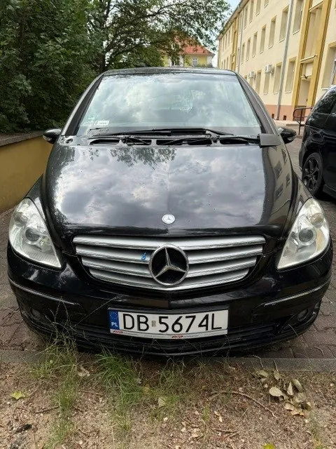 Czarny samochód marki Mercedes-Benz zaparkowany przodem do obiektywu. Na przodzie widoczny jest charakterystyczny grill z logo marki oraz tablica rejestracyjna o numerze DB 5674L. W tle widać zielone drzewa oraz żółty budynek.