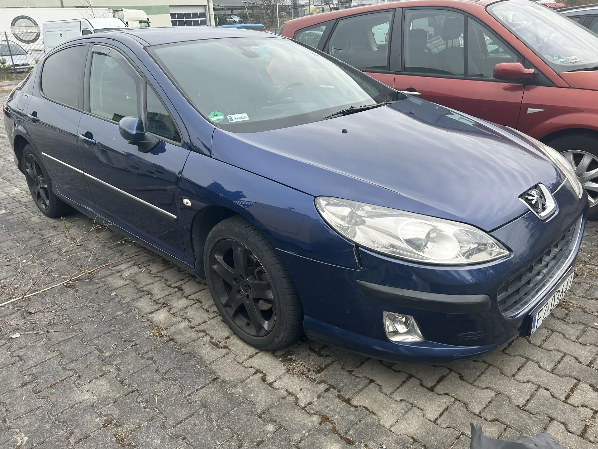 Niebieski samochód marki Peugeot z czarnymi felgami zaparkowany na brukowanym placu. Widoczna tablica rejestracyjna FZ 036 AM oraz naklejki na przedniej szybie. W tle czerwony samochód, budynek przemysłowy i inne pojazdy. Na ziemi sucha trawa i gałęzie.