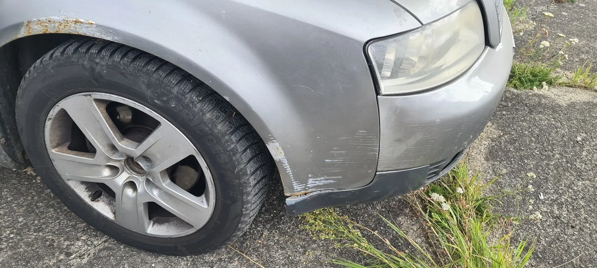 Zbliżenie na przedni lewy błotnik i koło srebrnego samochodu. Na błotniku, nad nadkolem i na zderzaku widoczne są uszkodzenia: rdza na górnej krawędzi oraz liczne zarysowania i odpryski lakieru w dolnej części. Widoczna jest srebrna felga aluminiowa z pięcioma ramionami i opona. Samochód stoi na asfaltowej nawierzchni z kępami trawy.