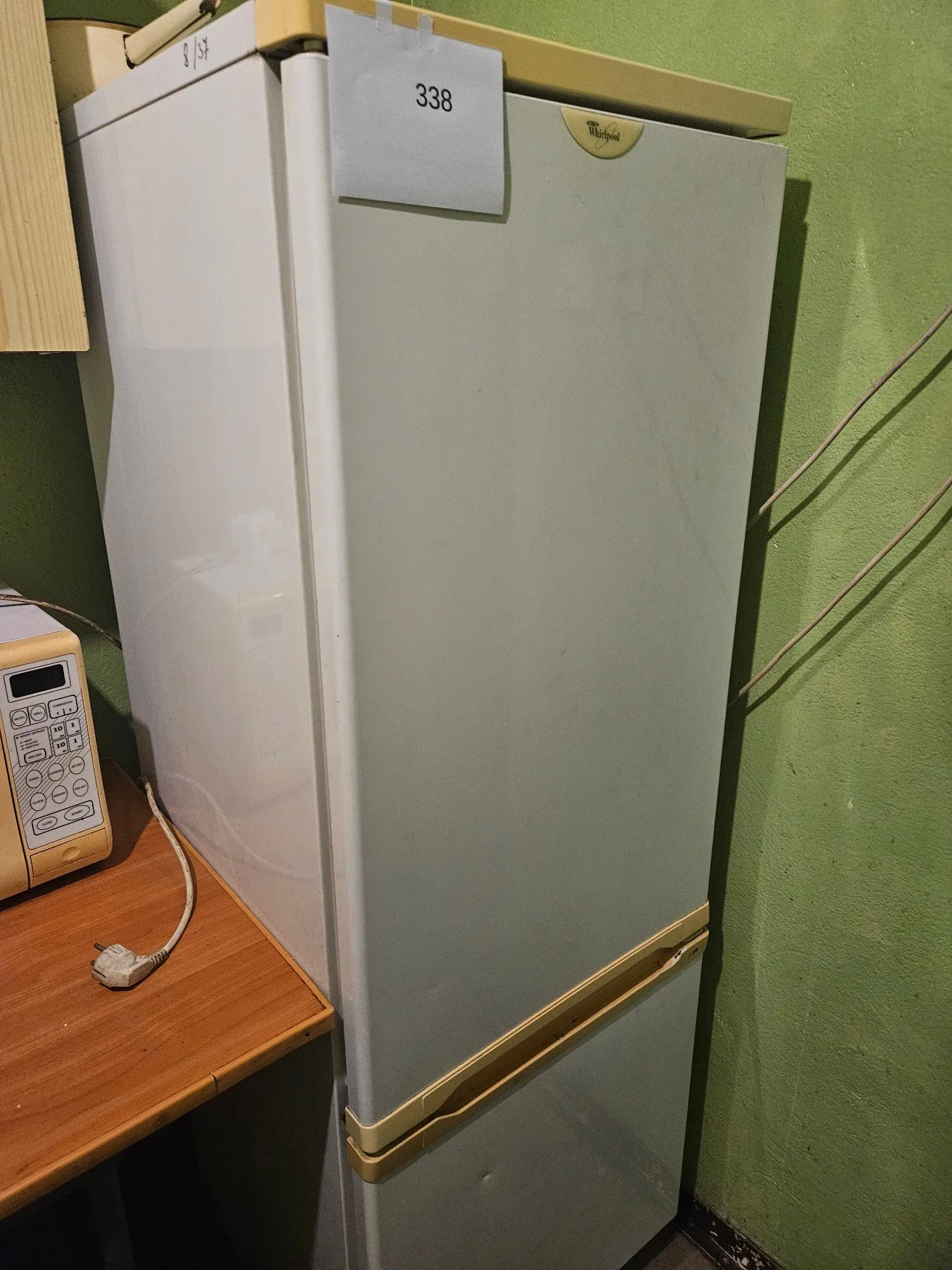 Biała lodówka dwudrzwiowa marki Whirlpool stojąca przy zielonej ścianie. Na górnej części lodówki przyklejona jest biała kartka z numerem 338. Po lewej stronie widoczny jest fragment drewnianego blatu, na którym stoi beżowy telefon stacjonarny z klawiaturą i wyświetlaczem oraz kabel z wtyczką.