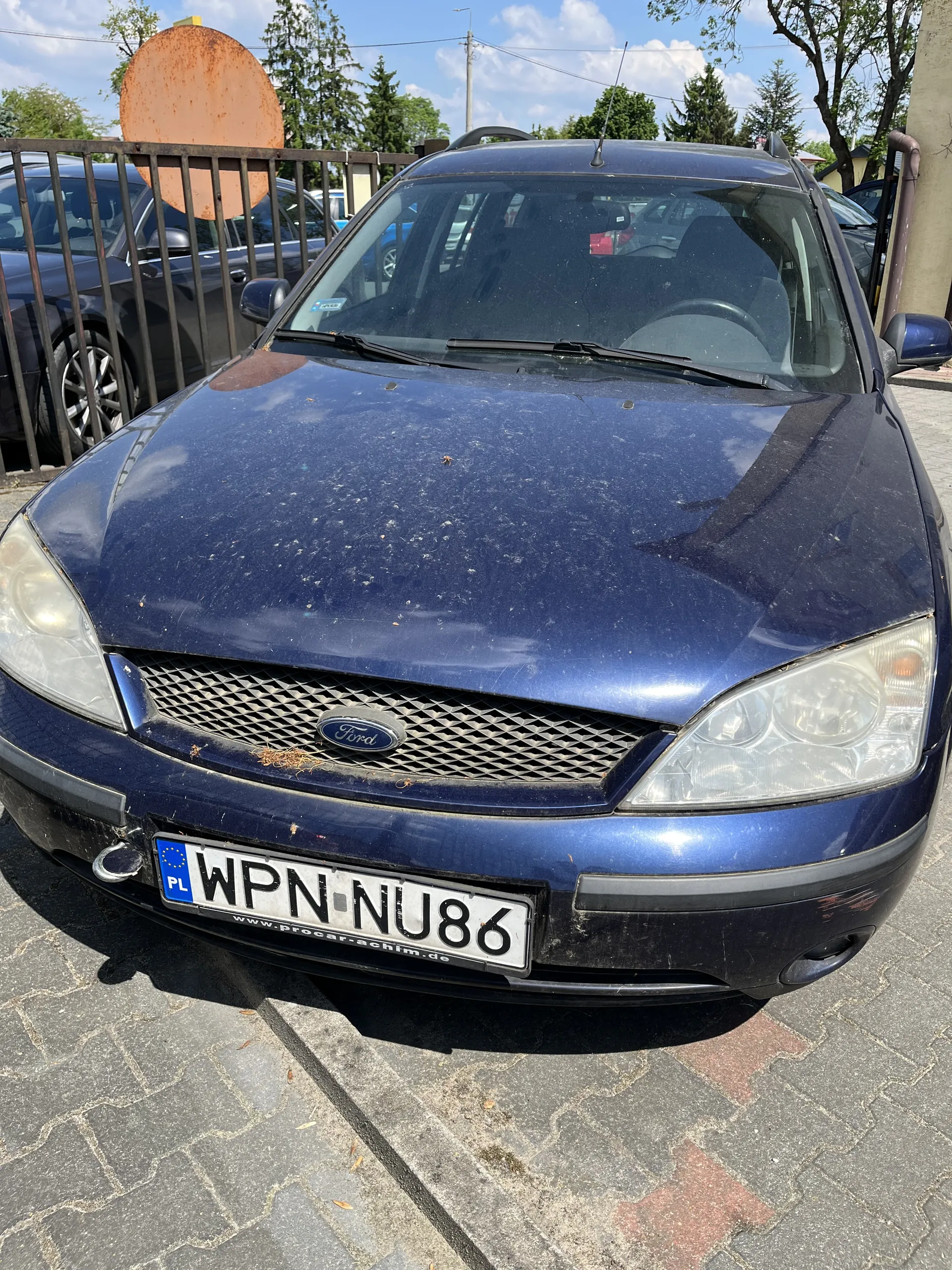 Niebieski samochód marki Ford z tablicą rejestracyjną WPN NU86 zaparkowany na brukowanej powierzchni. Widoczne są drzewa, ogrodzenie oraz inne pojazdy na tle. Niebo z chmurami. Na masce samochodu znajdują się ślady brudu.