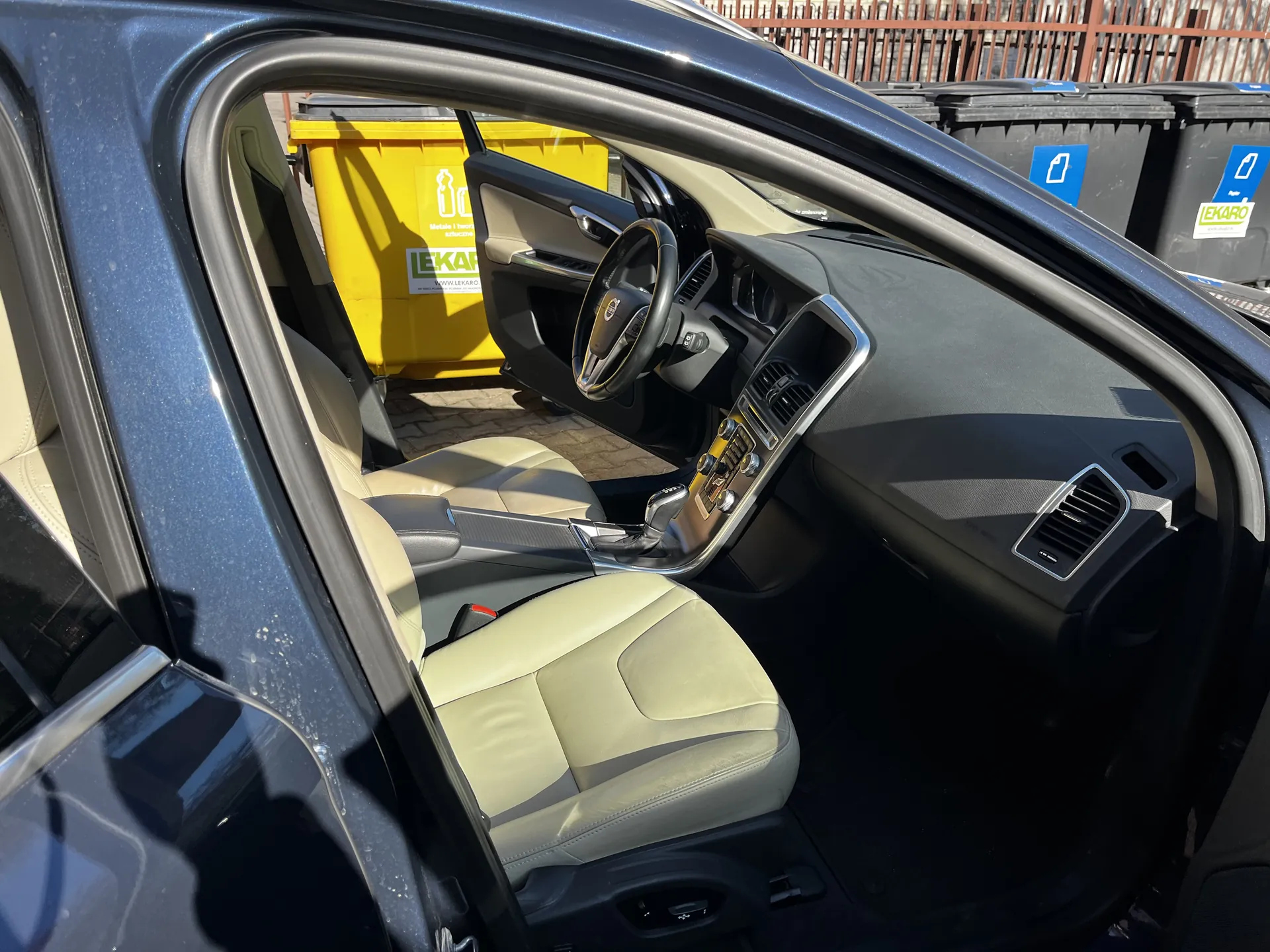 Wnętrze samochodu z otwartymi drzwiami od strony kierowcy. Widoczne są beżowe fotele, kierownica, deska rozdzielcza z przyciskami i gałką zmiany biegów. Na zewnątrz, za samochodem, stoją żółte i czarne pojemniki na odpady z napisem "LEKARO" oraz niebieskimi naklejkami. W tle metalowe ogrodzenie.