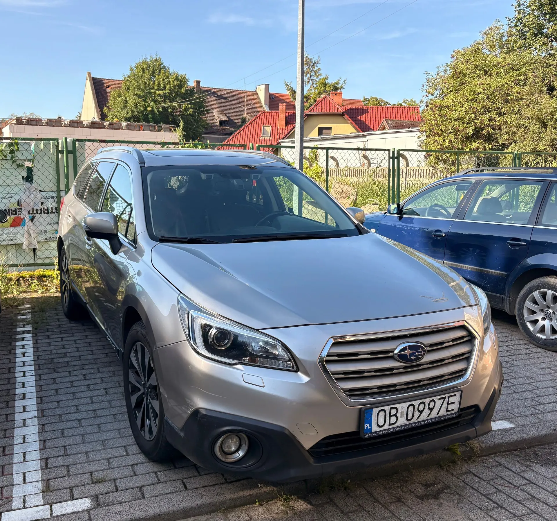 Srebrny samochód marki Subaru zaparkowany na kostce brukowej obok niebieskiego samochodu. W tle widoczny jest zielony płot, drzewa oraz domy z czerwonymi dachami pod jasnym, niebieskim niebem. Samochód ma polską tablicę rejestracyjną o numerze OB 0997E.