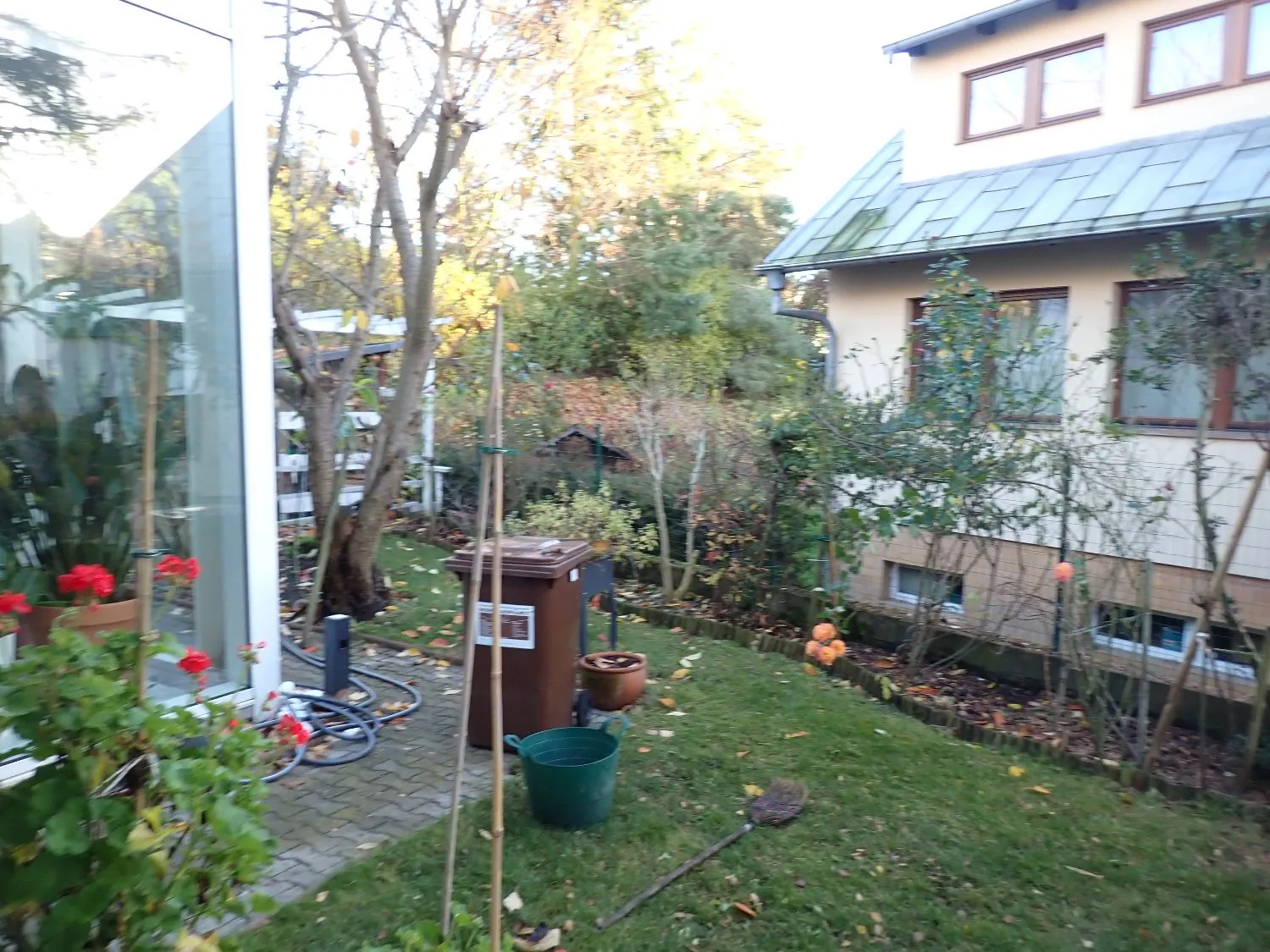 Widok na ogród przydomowy z fragmentem jasnego budynku po prawej stronie. Na trawniku stoi brązowy kompostownik, zielona donica oraz narzędzie ogrodnicze. Po lewej stronie widoczna jest szklana ściana oranżerii z roślinami i czerwonymi kwiatami. W tle znajdują się drzewa i krzewy w jesiennych barwach.