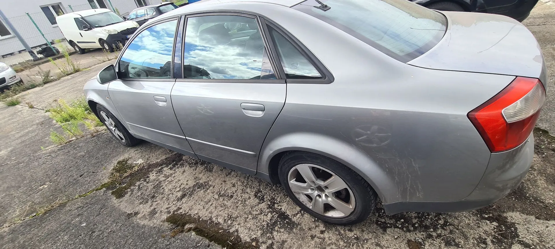 Srebrny samochód osobowy typu sedan zaparkowany na betonowym podłożu. Widok z boku i z tyłu pojazdu. W tle widoczny jest biały samochód dostawczy oraz fragment budynku.