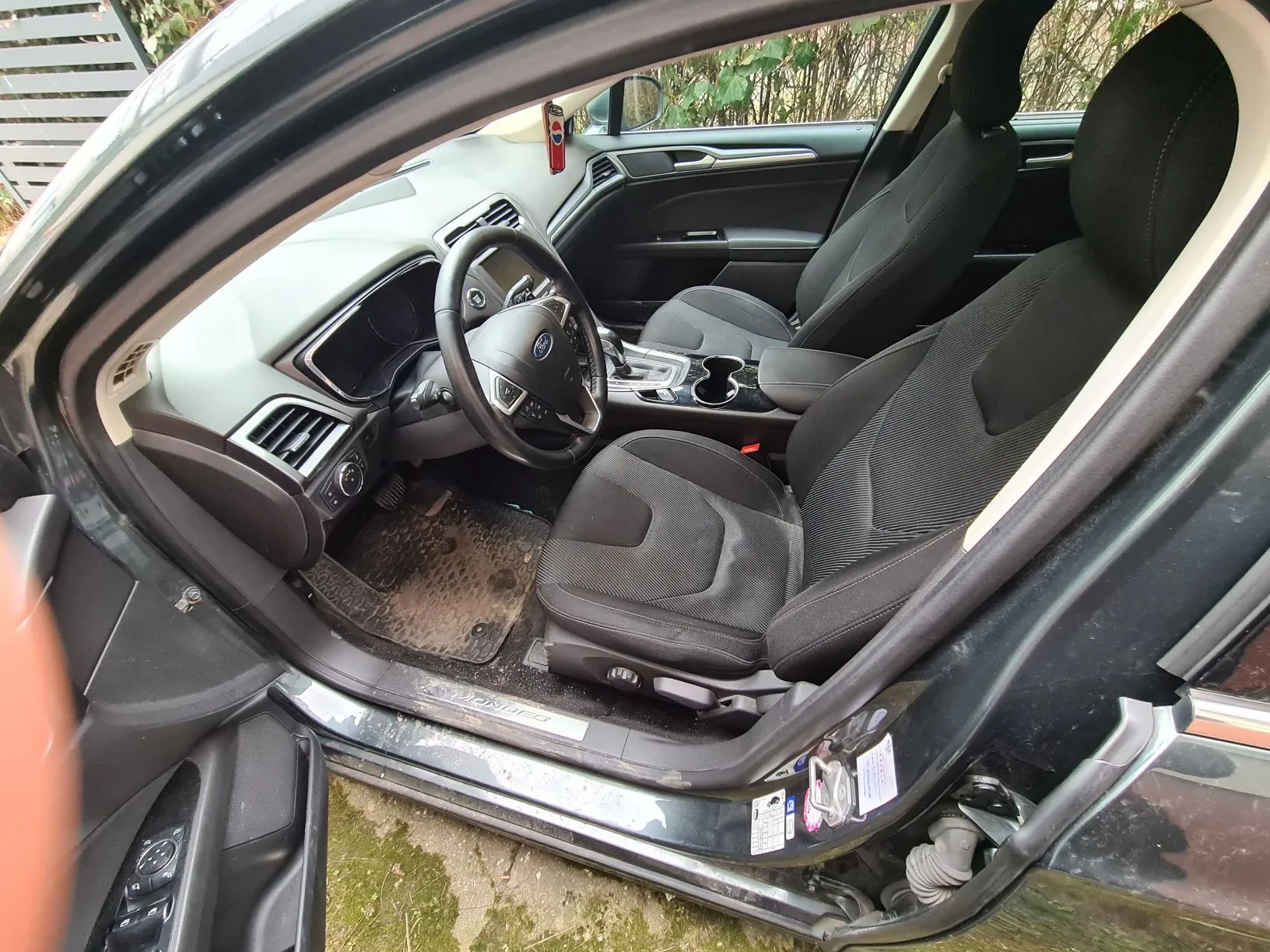 Wnętrze samochodu z otwartą przodem drzwiami od strony kierowcy. Widoczne czarne siedzenia, kierownica z logo Ford, deska rozdzielcza z wentylacjami i przyciskami, brudne dywaniki podłogowe. Na progu drzwi napis "MONDEO". W tle za szybą widoczne krzewy i metalowe ogrodzenie. Na lusterku wstecznym wiszący czerwony odświeżacz powietrza. Na drzwiach naklejki z dokumentacją techniczną.
