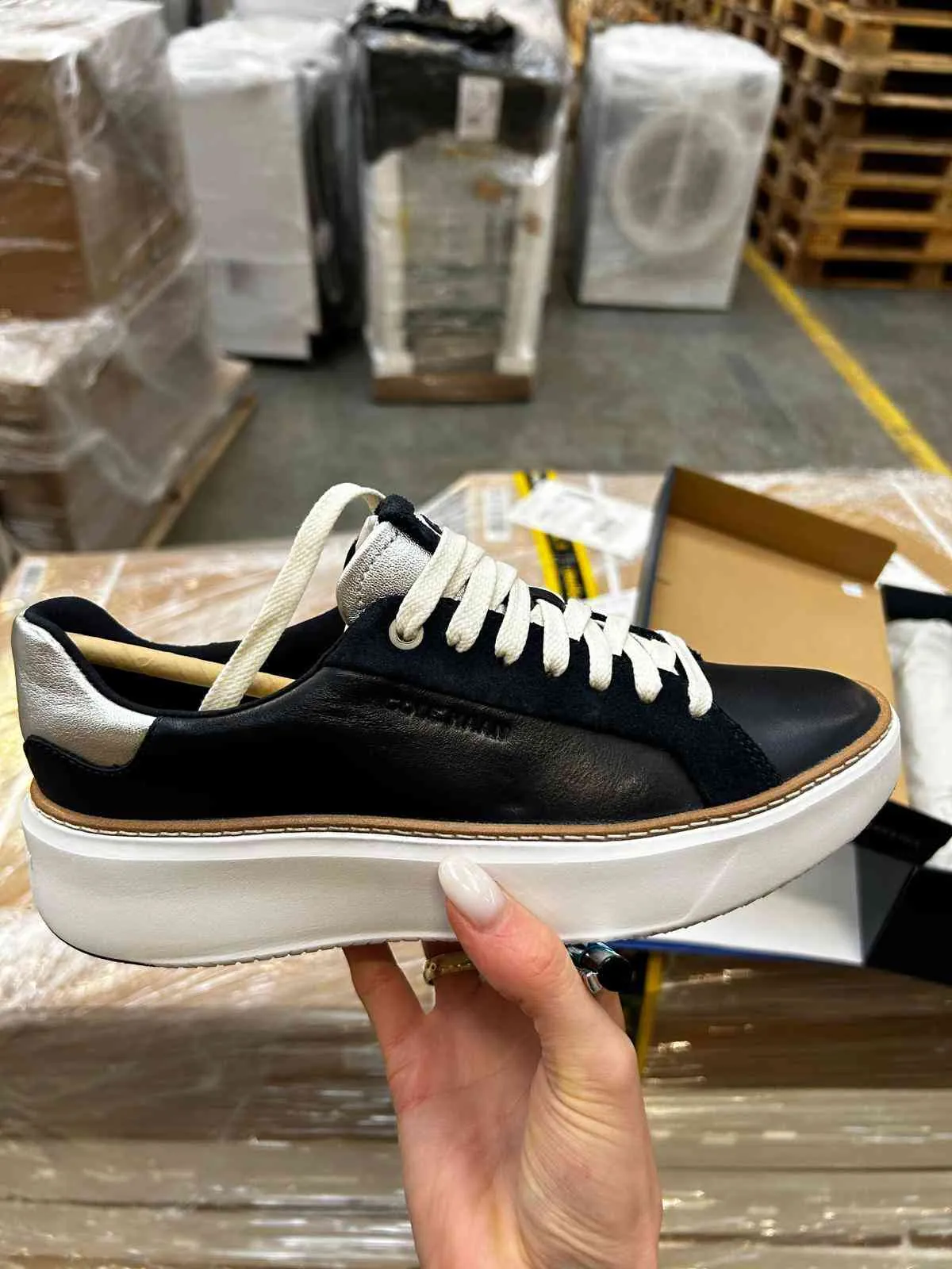 Ręka trzyma czarny trampki z białymi sznurówkami, białą podeszwą platformową i srebrnymi akcentami na pięcie. Na boku trampki widoczny jest napis "COPENHAGEN". Tło przedstawia magazyn z ułożonymi w stosy kartonami owiniętymi folią oraz drewnianymi paletami.