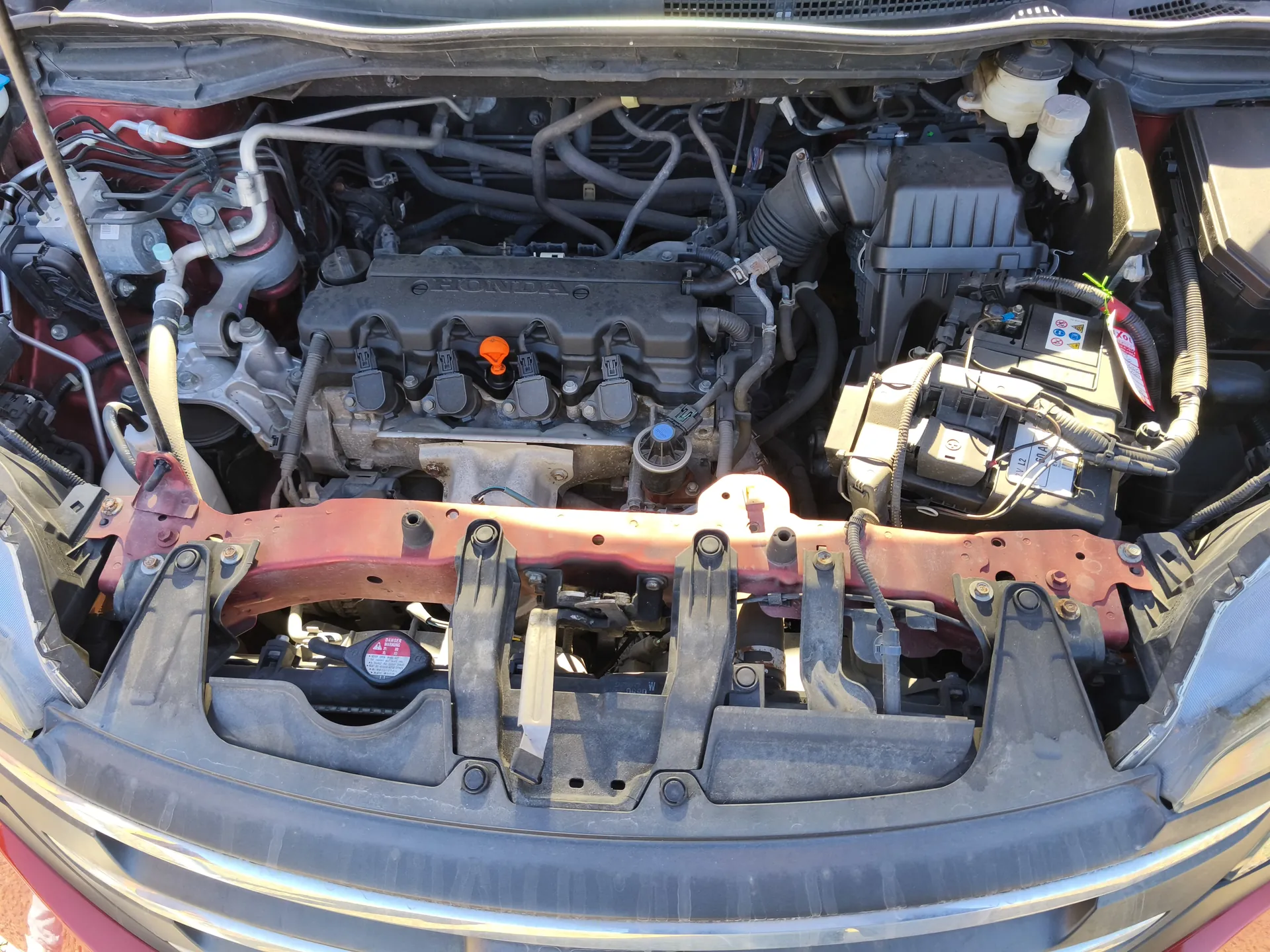 Widok otwartej maski samochodu z silnikiem marki HONDA. W centralnej części widoczna czarna pokrywa silnika z napisem "HONDA". Obecne są różne elementy: przewody, węże, czerwona belka podtrzymująca, zbiornik płynu chłodniczego z pomarańczową nakrętką oraz akumulator po prawej stronie z etykietami. Na przedniej części silnika znajduje się czerwony naklejony znak ostrzegawczy. Całość ukazana jest z góry, z widocznymi metalowymi i plastиковymi częściami.