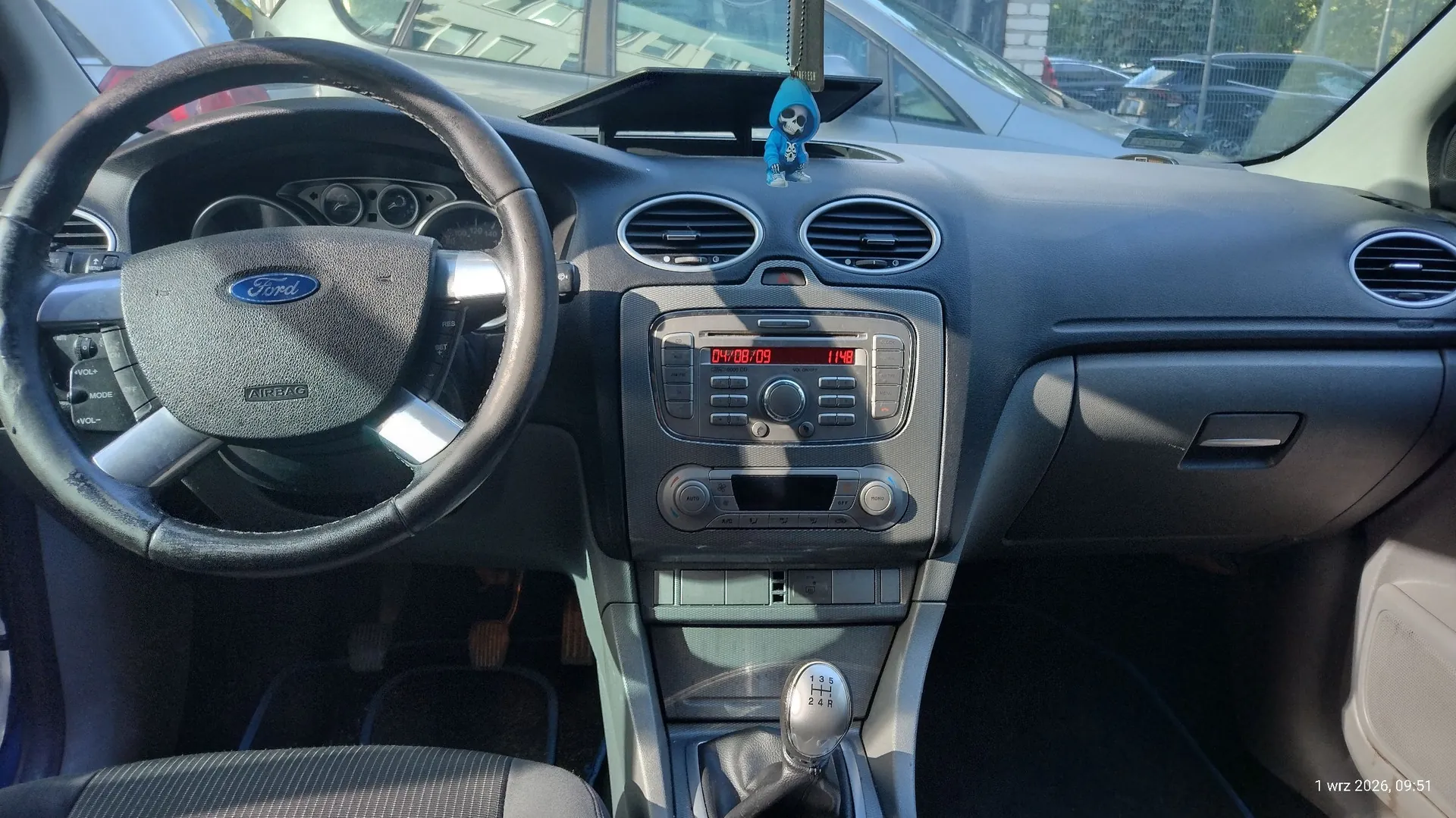Wnętrze samochodu marki Ford widziane z perspektywy kierowcy. Widoczna jest czarna kierownica z logo Ford, deska rozdzielcza z centralnym radiem wyświetlającym czerwony tekst '04/08/09 11:48', nawiewy powietrza oraz lewarek zmiany biegów. Na górnej części deski rozdzielczej znajduje się mała niebieska figurka. W prawym dolnym rogu widnieje data i godzina '1 wrz 2026, 09:51'.