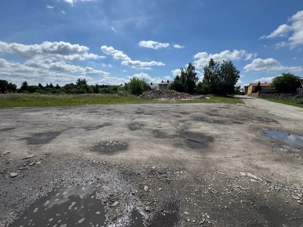 Szeroki widok na zniszczony, asfaltowy plac z licznymi dziurami i kałużami wody. W tle znajduje się pryzma gruzu, zielone krzewy, drzewa oraz kilka niskich budynków mieszkalnych pod jasnoniebieskim niebem z rozproszonymi białymi chmurami.