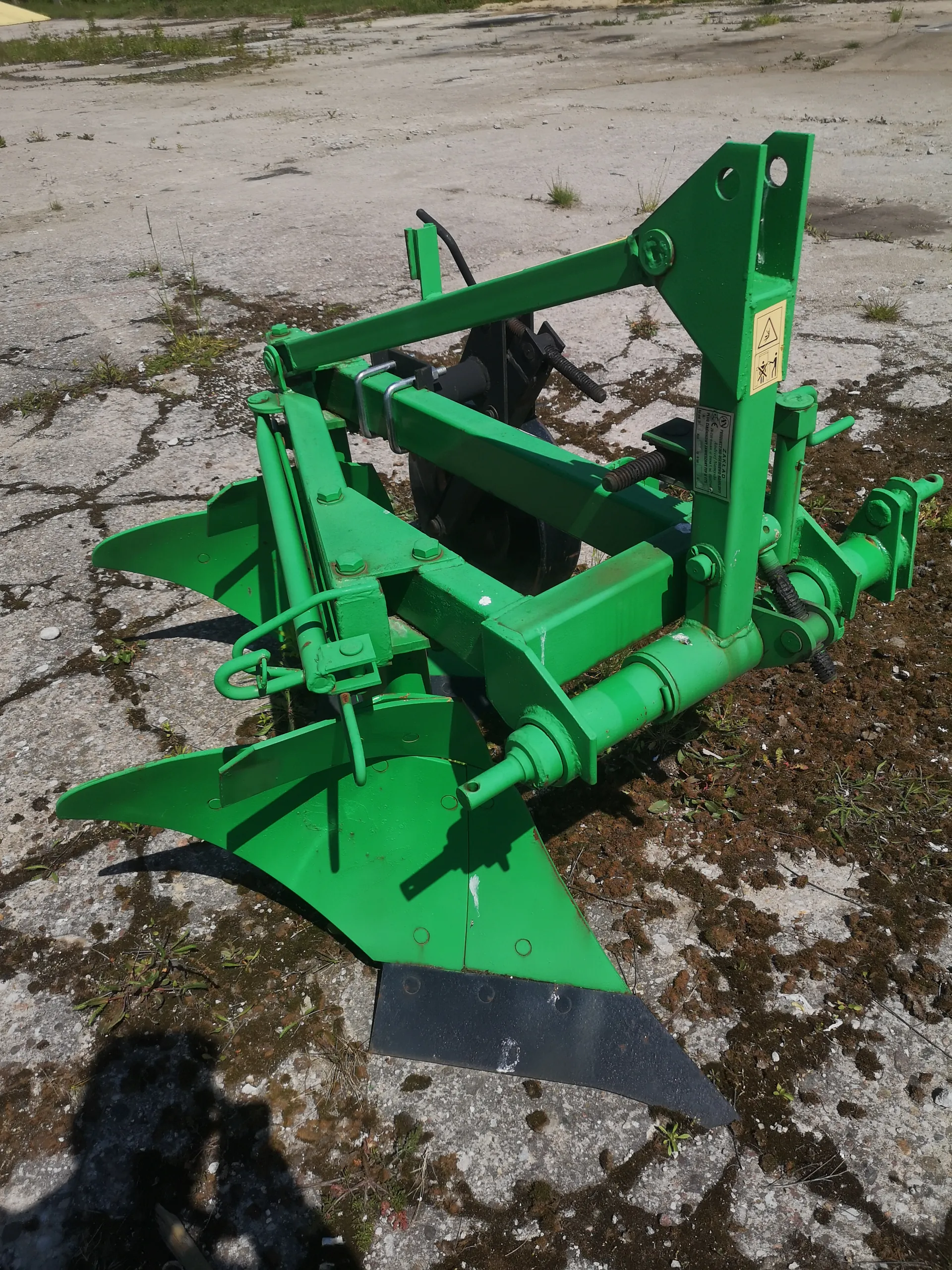 Zielony metalowy sprzęt rolniczy, prawdopodobnie pług lub kultywator, z widocznymi ostrzami i mechanizmami mocującymi. Urządzenie stoi na pękniętej betonowej powierzchni z rozproszoną trawą i mchem. W tle otwarta przestrzeń z betonem i rzadką roślinnością. Na maszynie znajduje się żółty znak ostrzegawczy z symbolami oraz metalowa tabliczka z nieczytelnym tekstem.
