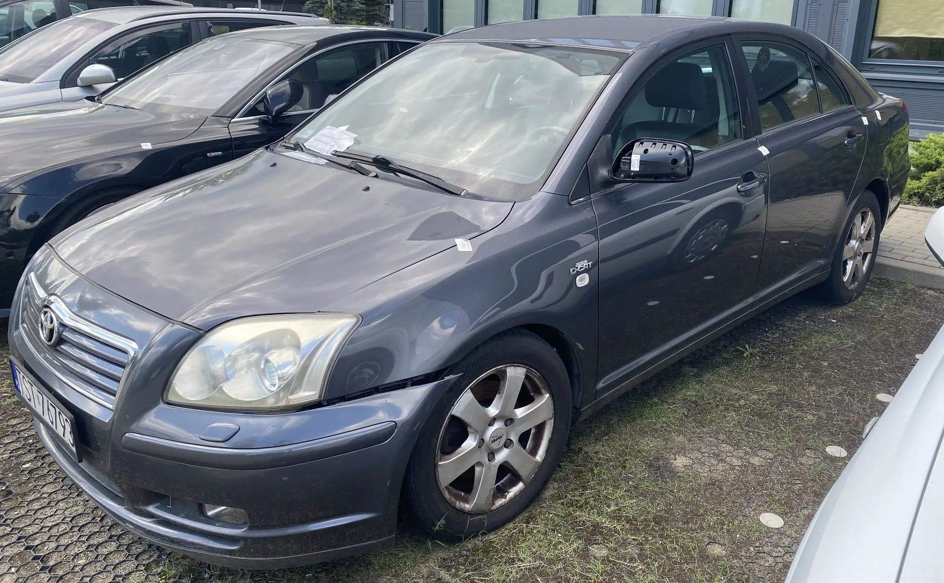Szary samochód osobowy marki Toyota zaparkowany na placu z kostki brukowej i trawy. Pojazd jest widoczny z przodu i z boku, z lekkim uszkodzeniem zderzaka przedniego po stronie kierowcy. W tle widoczne są inne zaparkowane samochody oraz fragment budynku.
