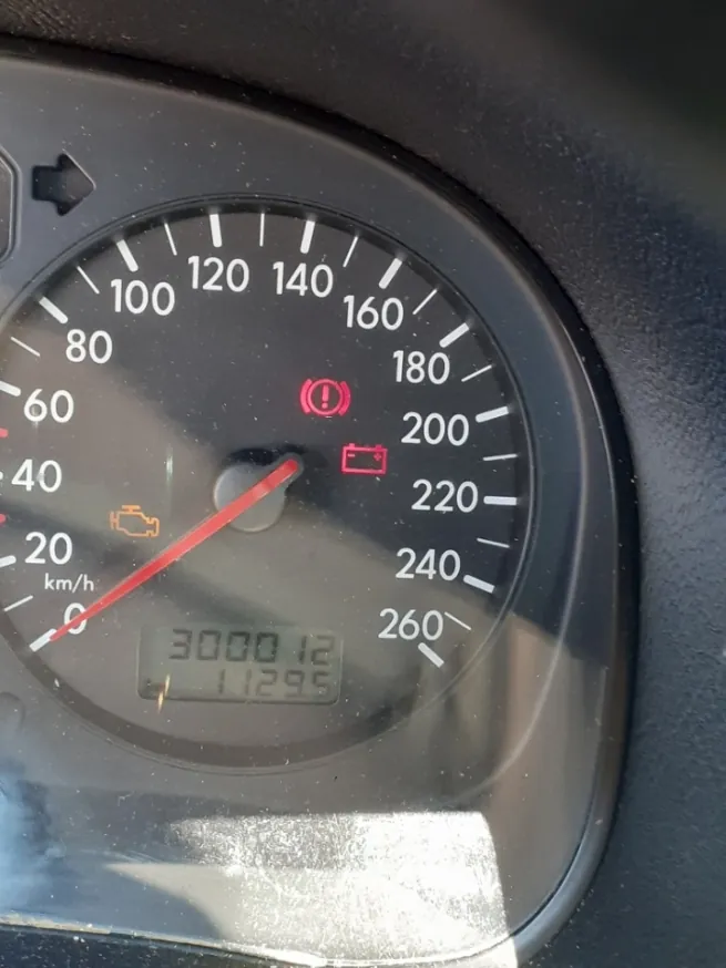 Zbliżenie na prędkościomierz w samochodzie. Tarcza z podziałką od 0 do 260 km/h z czerwoną wskazówką wskazującą wartość bliską 0. Na tarczy widoczne są zapalone kontrolki ostrzegawcze: pomarańczowa ikona silnika, czerwona ikona układu hamulcowego oraz czerwona ikona akumulatora. W dolnej części znajduje się cyfrowy licznik przebiegu wyświetlający liczby 3000 12 oraz 11295.
