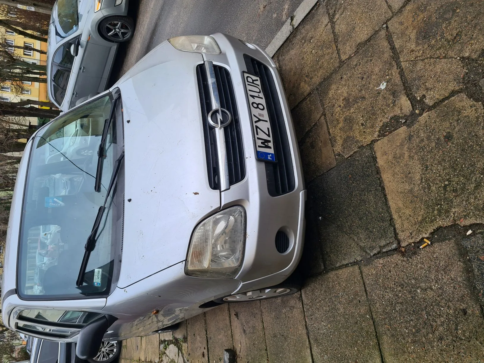 Srebrny samochód zaparkowany przy chodniku, widok z przodu. Tablica rejestracyjna z napisem WZY 81UR oraz niebieskim pasem z oznaczeniem PL. W tle drugi samochód, budynki i drzewa. Chodnik z kamiennymi płytami po prawej stronie obrazu.