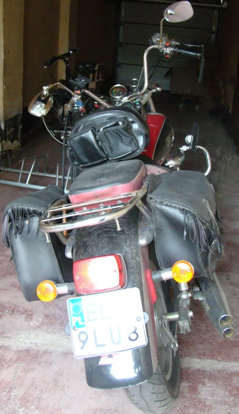 Widok z tyłu na czerwony motocykl zaparkowany w garażu. Motocykl posiada czarne skórzane sakwy po obu stronach, czarne oparcie dla pasażera oraz chromowany bagażnik. Widoczna jest tablica rejestracyjna z napisem „9LU8” oraz czerwone światło stopu i dwa żółte kierunkowskazy. W tle widoczny jest drugi motocykl.