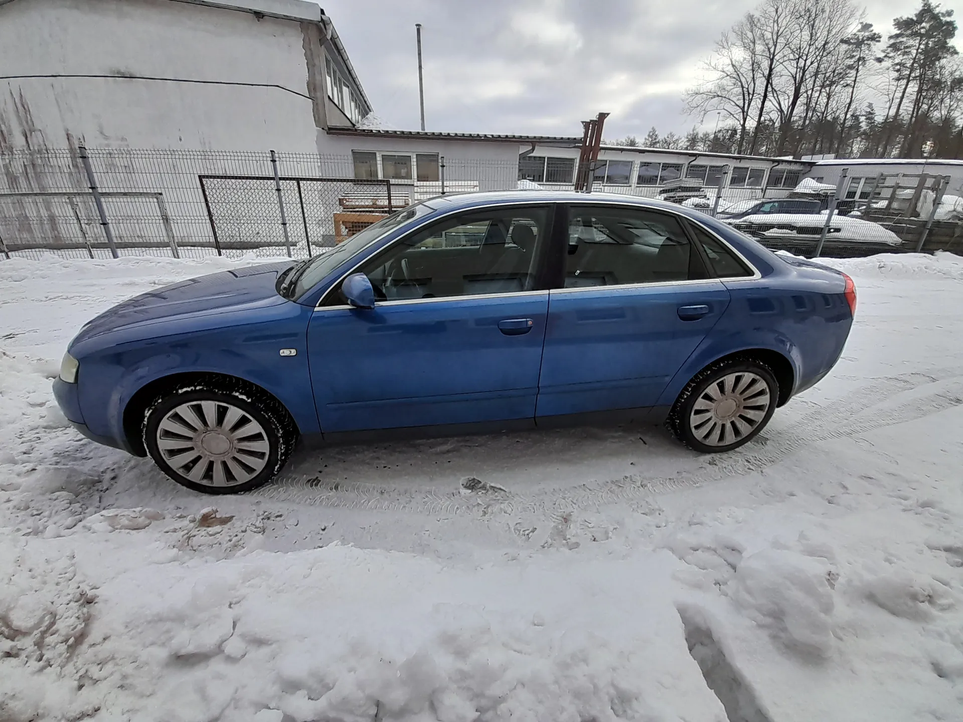 Niebieski czterodrzwiowy sedan Audi zaparkowany na śnieżnej powierzchni. W tle białe budynki z oknami, metalowe ogrodzenie siatkowe i drzewa bez liści. Niebo zachmurzone, opony zimowe na srebrnych felgach. Śnieg pokrywa ziemię i częściowo nadwozie.