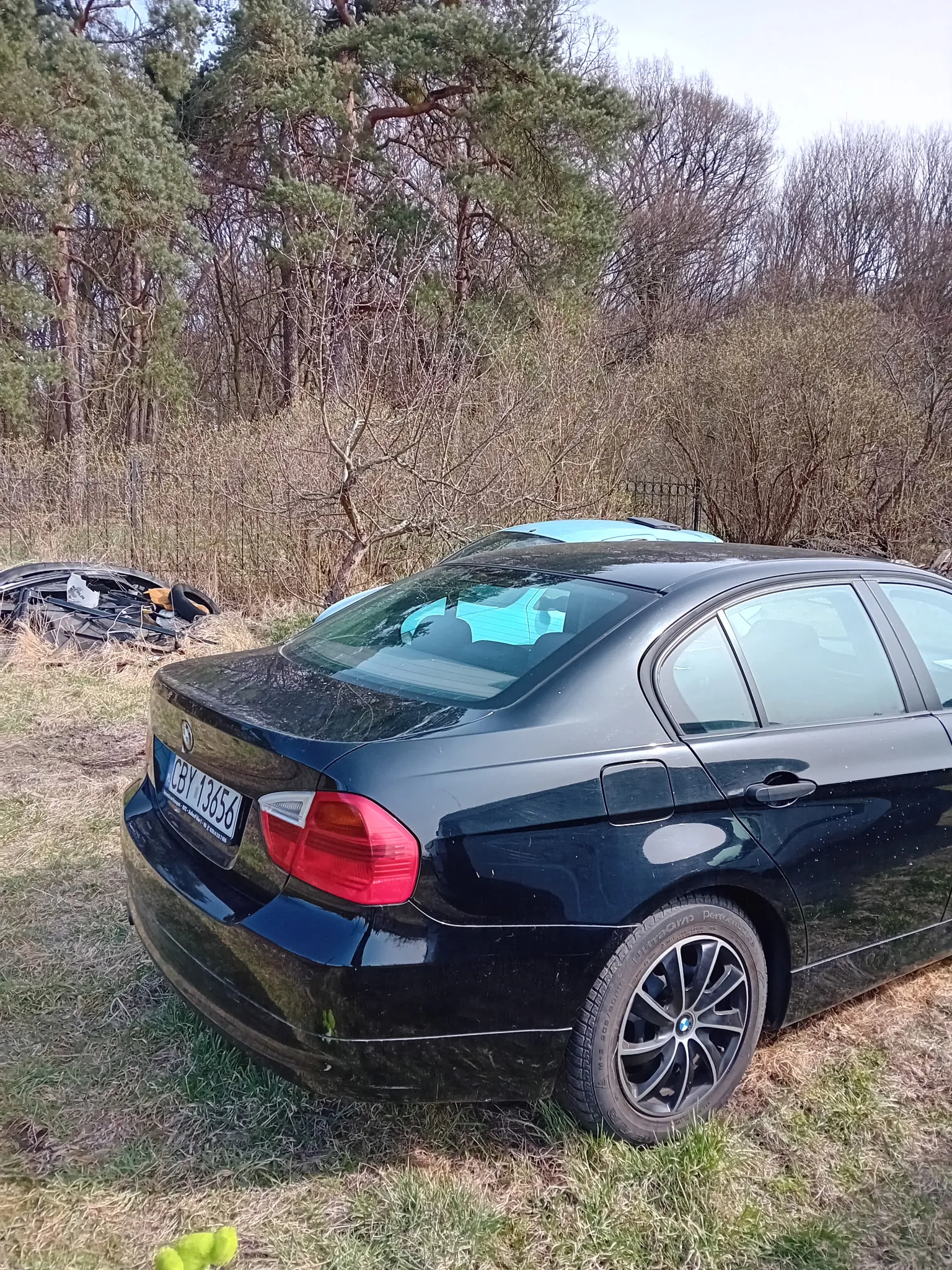 Czarny samochód marki BMW, model sedan, zaparkowany na trawie. Widoczny tył pojazdu z tablicą rejestracyjną CBY 13656. W tle drzewa, krzewy oraz częściowo widoczny niebieski samochód. Po lewej stronie obrazu stos zużytych opon i innych elementów. Trawa na terenie jest wysuszona, a drzewa mają mieszane liście i nagie gałęzie.