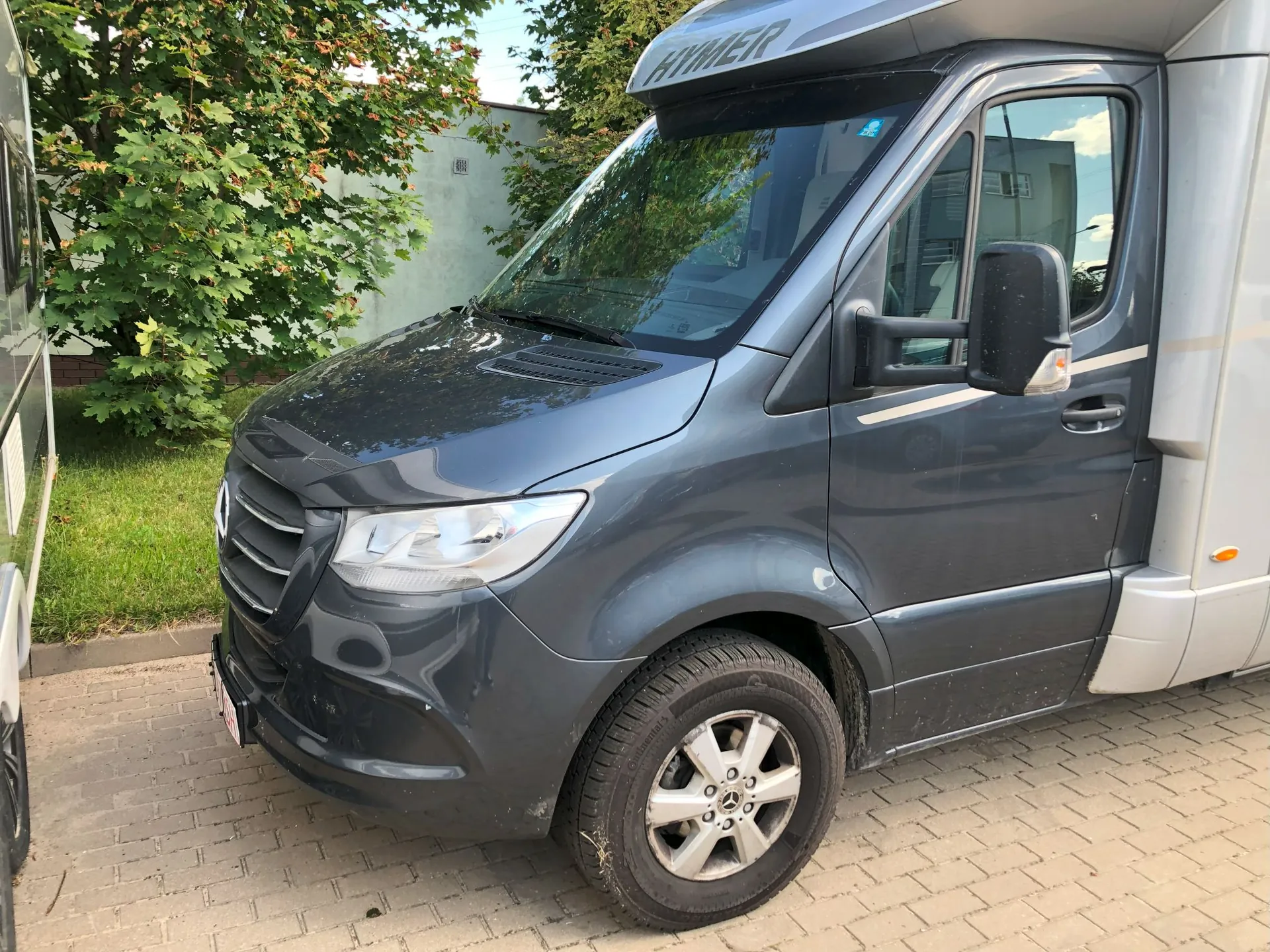Szary kamper marki Hymer zaparkowany na kostce brukowej. Widoczny jest przód i prawy bok pojazdu. Nad przednią szybą znajduje się napis 'HYMER'. W tle widać zielone drzewa i fragment jasnej ściany budynku.
