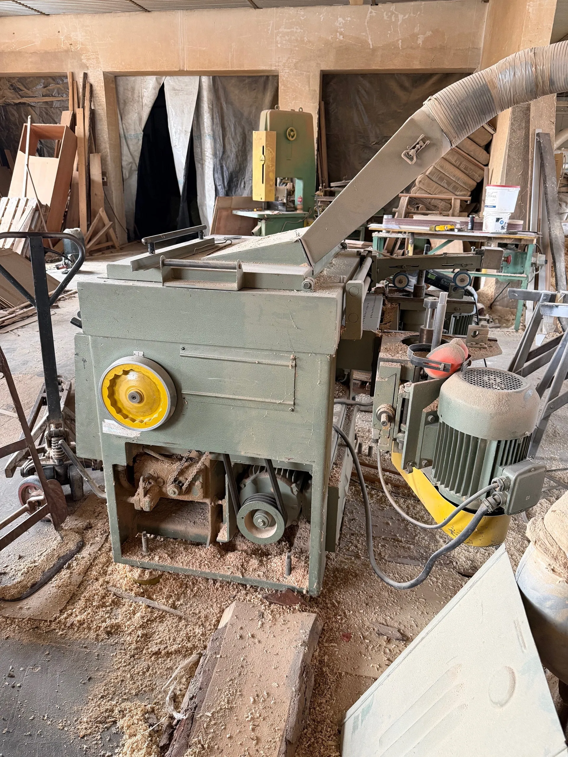 Wnętrze warsztatu stolarskiego z zieloną maszyną przemysłową o żółtym kole. Na podłodze widoczne są wióry drewna, a w tle znajdują się inne urządzenia, takie jak piła taśmowa, deski i materiały. Ściany pomieszczenia są beżowe, a na podłodze leżą narzędzia i elementy konstrukcyjne.