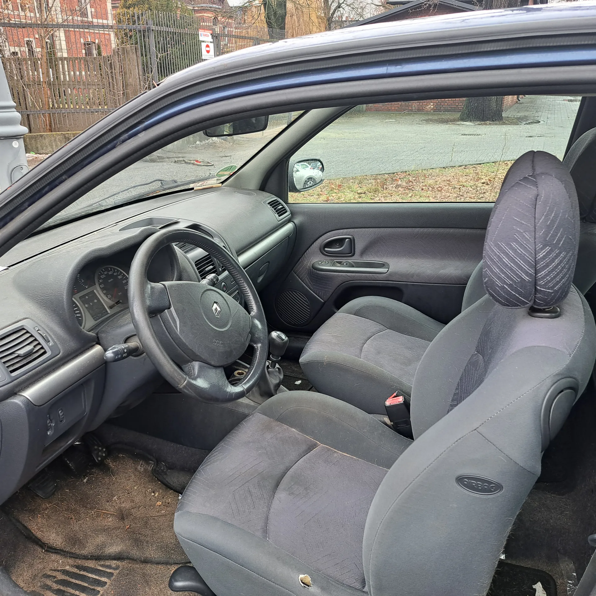 Wnętrze samochodu z widoczną kierownicą z logo Renault, deska rozdzielcza z licznikami, szare siedzenia z uszkodzeniem na siedzeniu kierowcy, drzwi z uchwytem i sterowaniem oknem. Poza samochodem widoczna ulica, ogrodzenie, czerwony znak zakazu wjazdu oraz budynki. Podłoga w samochodzie zanieczyszczona.