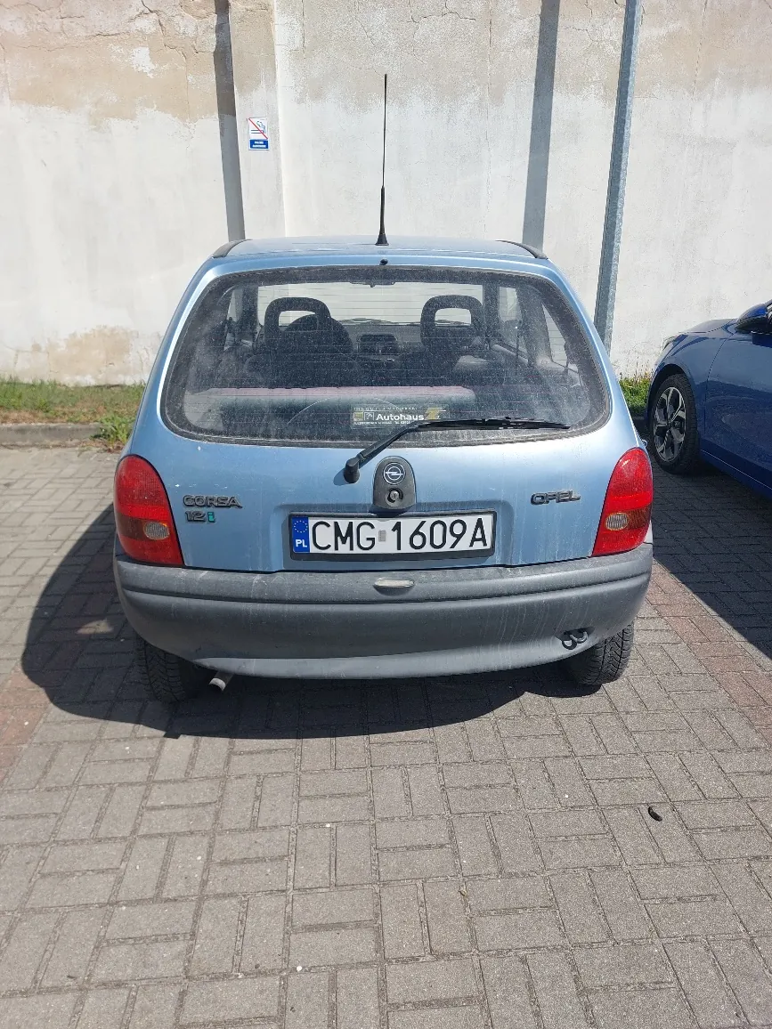 Z tyłu widoczny jest jasnoniebieski samochód Opel Corsa 12i z tablicą rejestracyjną CMG 1609A (kod kraju PL). Na tylnej szybie naklejka z napisem Autohaus. Samochód stoi na brukowanej powierzchni, w tle betonowa ściana z niebieskim znakiem zakazu. Po prawej stronie częściowo widoczny niebieski samochód. Tylne światła czerwone, zderzak szary, antena na dachu.