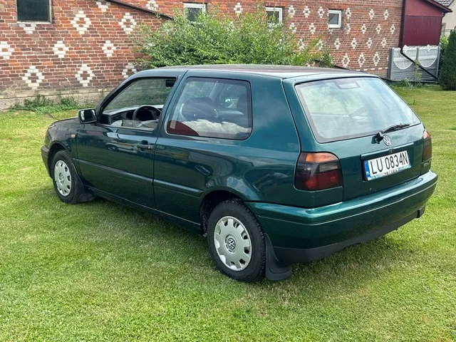 Ciemnozielony samochód Volkswagen Golf trzeciej generacji zaparkowany na trawie na tle ceglanego budynku z białymi zdobieniami. Widoczny tył i bok auta oraz polska tablica rejestracyjna o numerze LU 0834N.