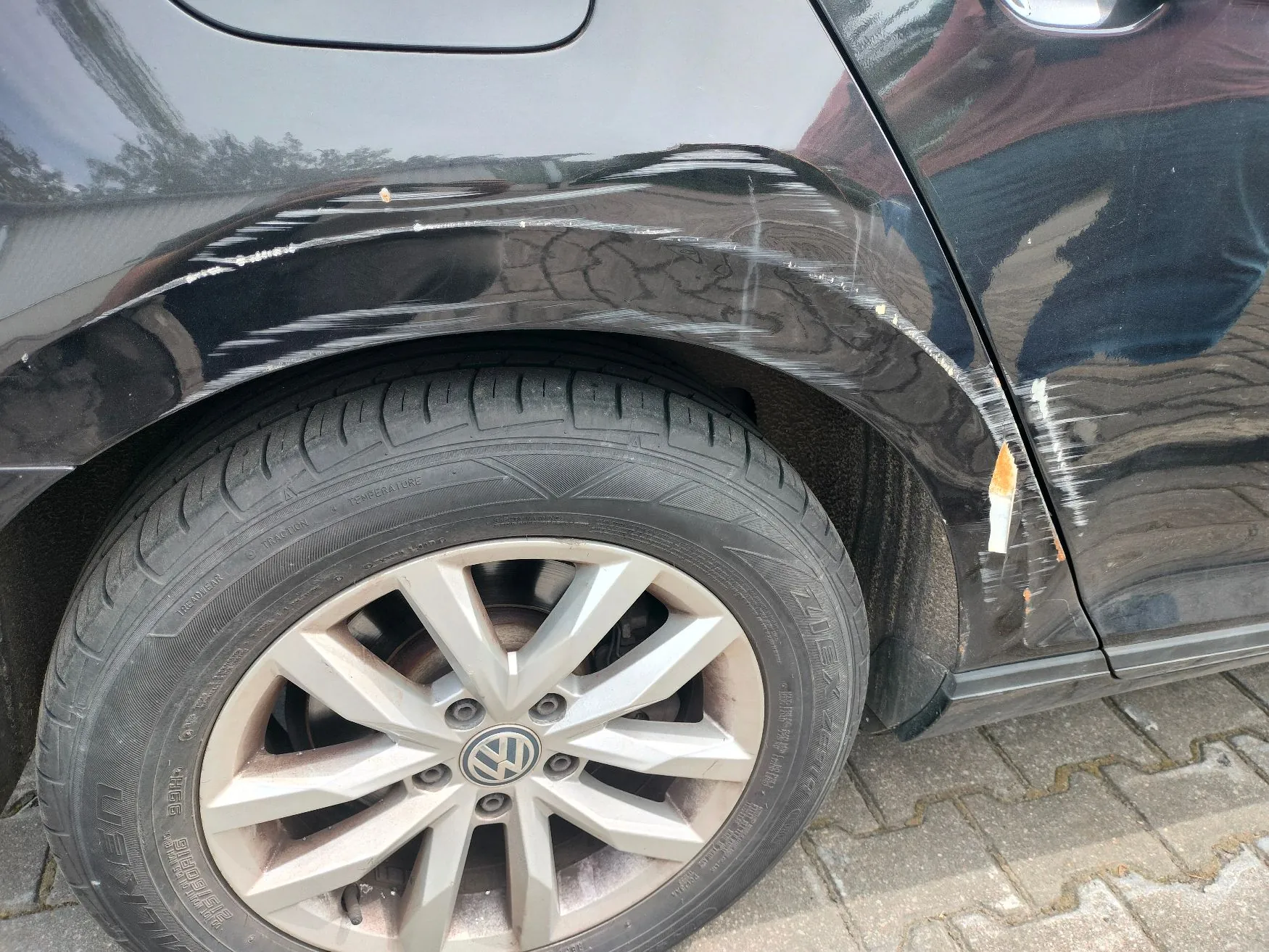 Zbliżenie na tylny lewy błotnik i koło czarnego samochodu. Na lakierze nad nadkolem widoczne są liczne, poziome zarysowania oraz wgniecenia. Koło posiada srebrną felgę aluminiową z logo Volkswagena w centrum oraz oponę zimową. Samochód stoi na kostce brukowej.