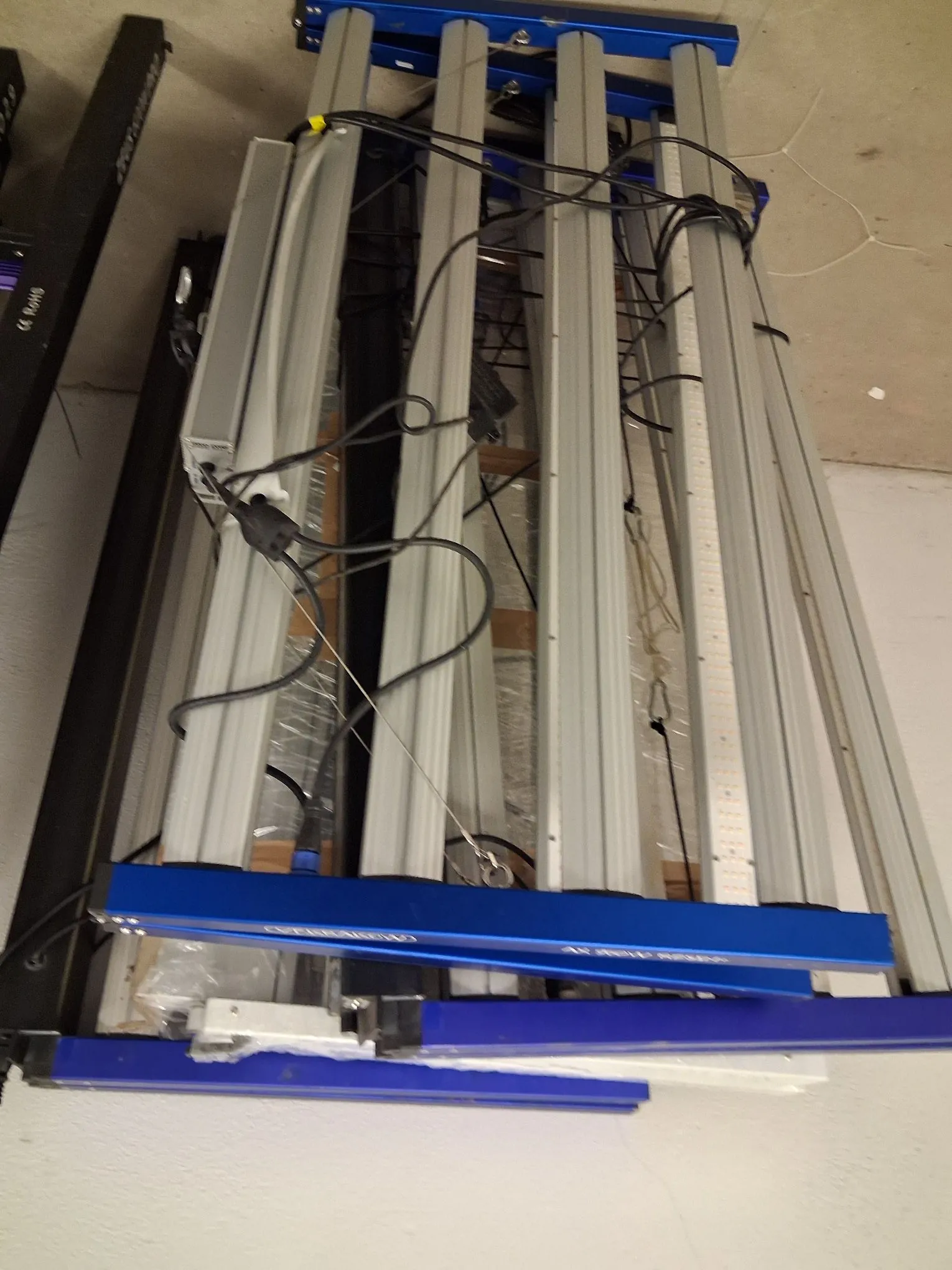 Zdjęcie przedstawiające konstrukcję z niebieskich profili aluminiowych, w której zamontowano kilka poziomych rzędów lamp LED w srebrnych oprawach. Widoczne są liczne czarne kable zasilające oraz elementy mocujące. Całość znajduje się w pomieszczeniu o białych ścianach.