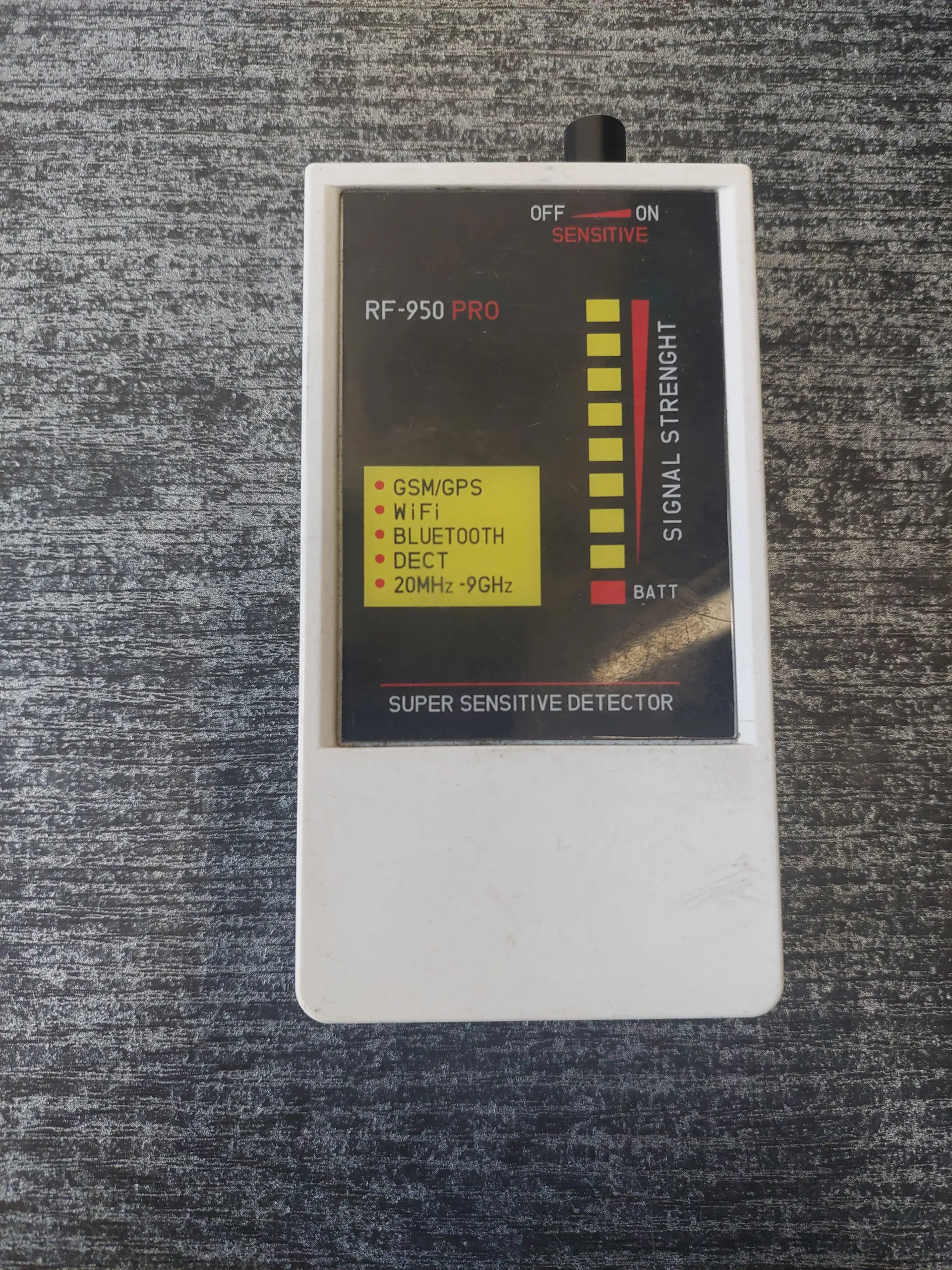 Biały prostokątny urządzenie oznaczone jako RF-950 PRO z czarnym wyświetlaczem. Na wyświetlaczu widoczny napis SIGNAL STRENGTH z czerwoną linią i żółtymi kwadratami wskazującymi poziom sygnału. Obok znajduje się wskaźnik BATT z czerwonym kwadratem. Po lewej stronie przełącznik z pozycjami OFF, ON i SENSITIVE. W żółtym polu listy: GSM/GPS, WiFi, BLUETOOTH, DECT, 20MHz-9GHz. Po prawej stronie wyświetlacza pionowy napis SUPER SENSITIVE DETECTOR. Urządzenie leży na szarej, teksturowanej powierzchni.