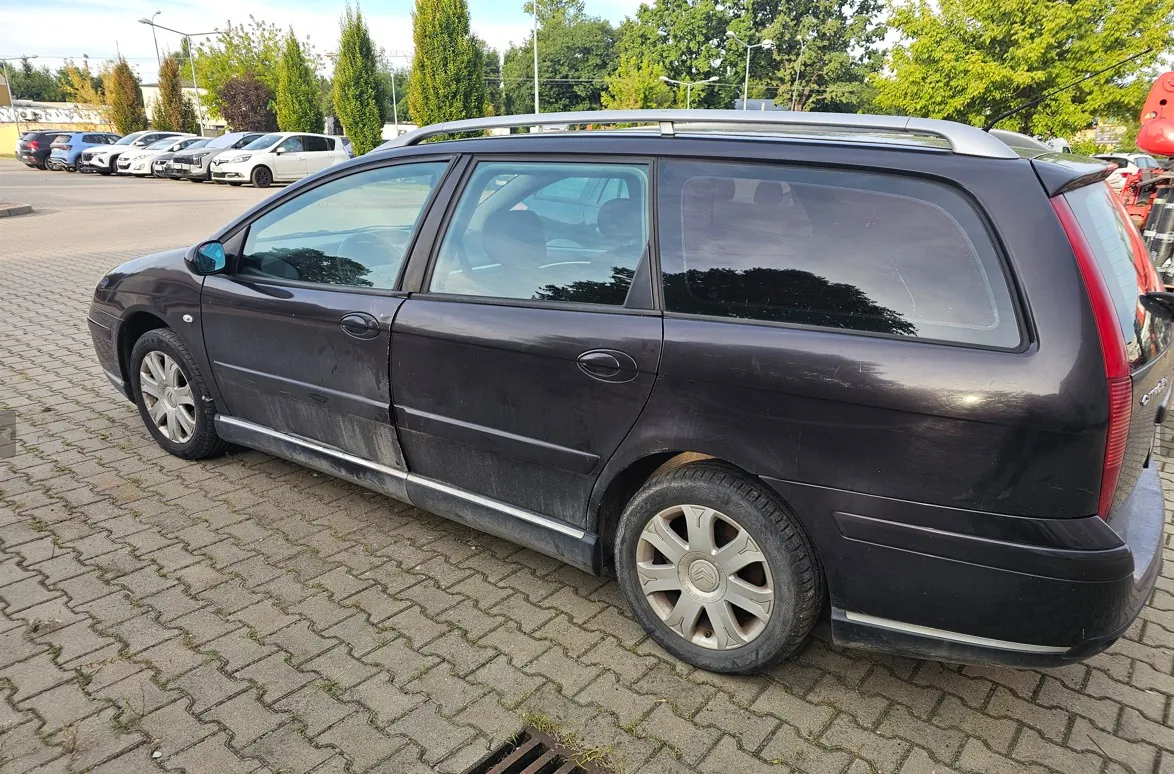 Czarny samochód typu kombi zaparkowany na kostce brukowej. Pojazd jest widoczny z profilu, posiada srebrne relingi dachowe i aluminiowe felgi. W tle widać inne zaparkowane samochody oraz zielone drzewa pod jasnym niebem.