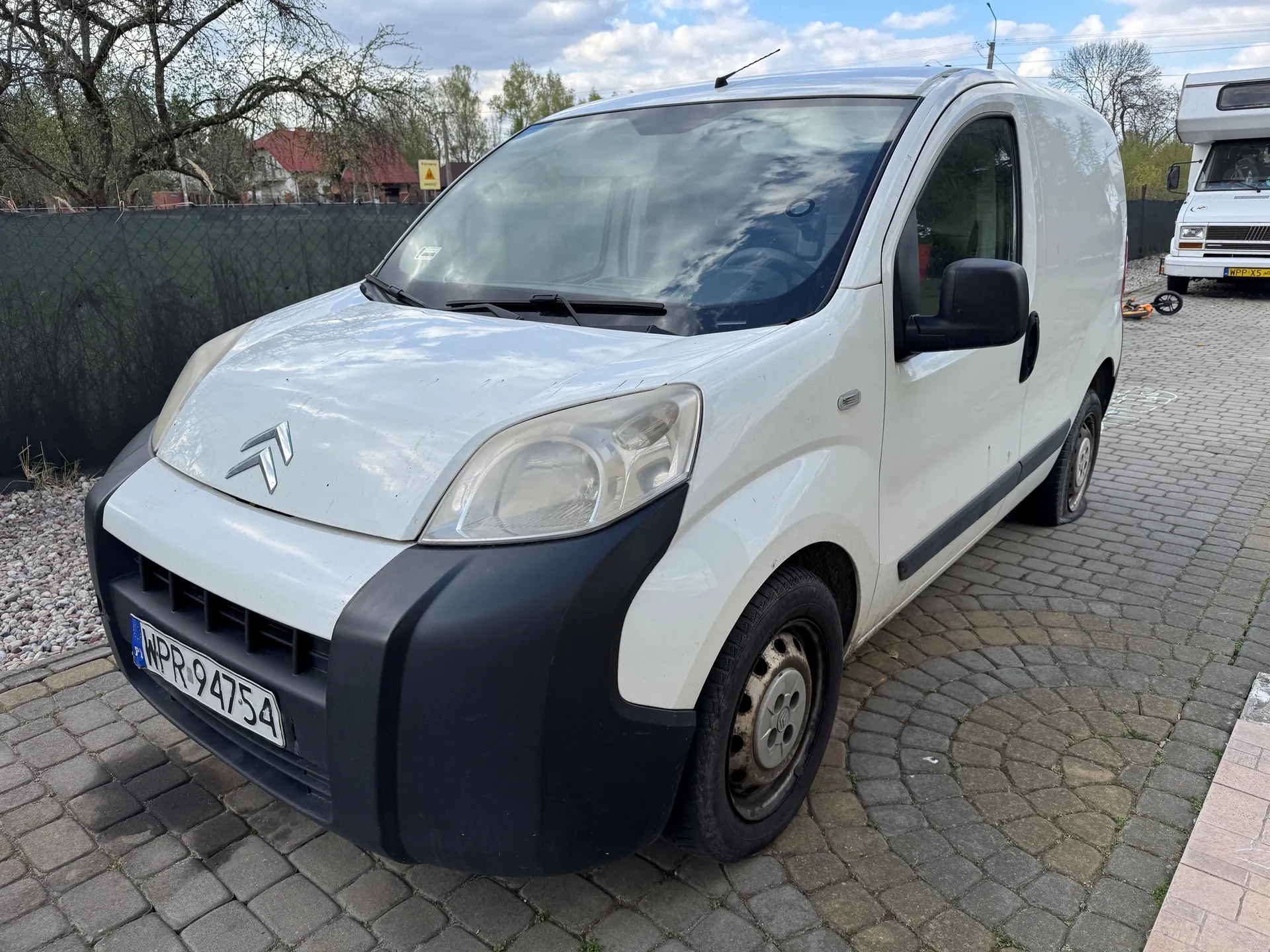 Biały samochód dostawczy Citroën z tablicą rejestracyjną WPR 94754 zaparkowany na brukowanej powierzchni. W tle widoczny biały pojazd typu camper, ogrodzenie, drzewa, dom z czerwonym dachem oraz niebo z chmurami. Na przedniej szybie odbijają się chmury i drzewa. Po prawej stronie obrazu znajduje się fragment pomarańczowego rowerka dziecięcego.