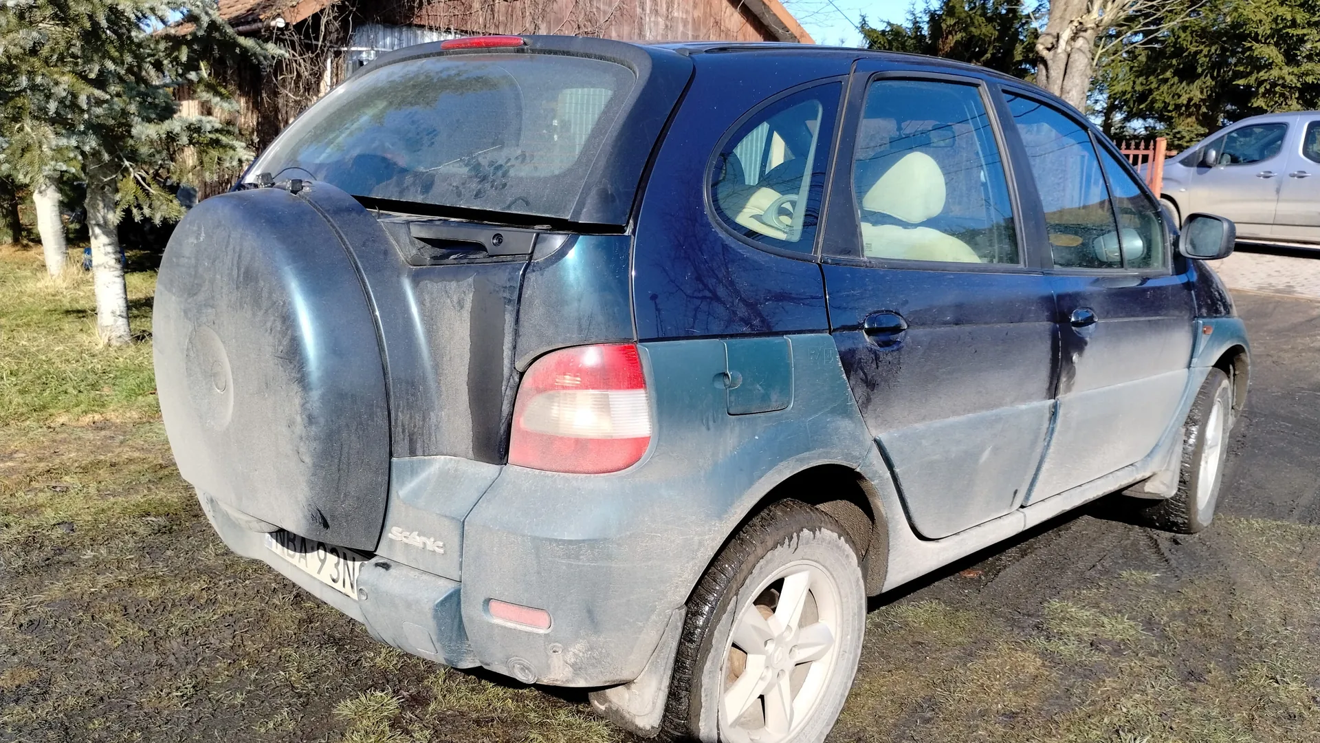 Niebieski samochód osobowy marki Dacia Sandero z zanieczyszczonym nadwoziem, widok z tyłu i boku. Na tylnej szybie widoczny czerwony reflektor, a na bagażniku osłona koła zapasowego. Tablica rejestracyjna z napisem MBA 93N. W tle drewniany budynek, drzewa iglaste, trawa oraz srebrny samochód zaparkowany na asfalcie. Samochód stoi na zanieczyszczonej trawie z widocznymi plamami błota.