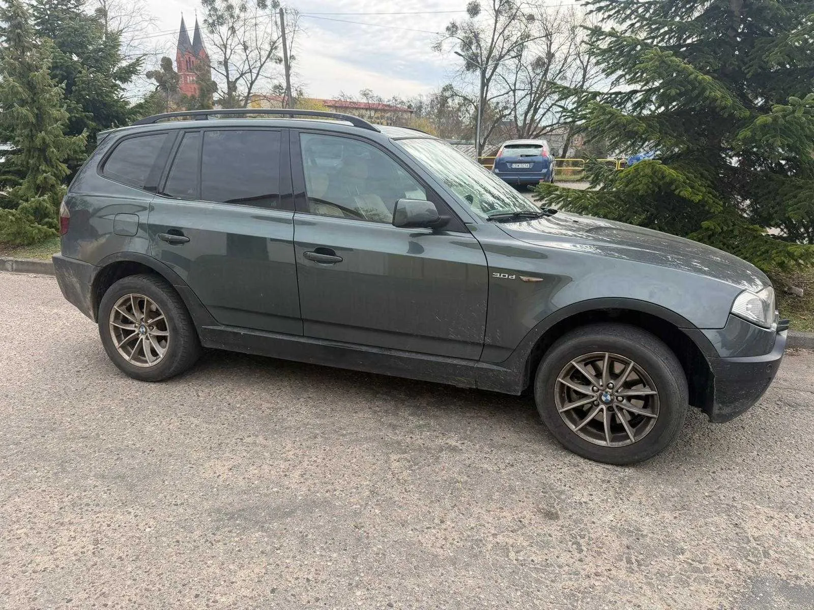 Ciemnoszary SUV marki BMW model 3.0d z dachowymi prętami i przyciemnianymi szybami zaparkowany na żwirowym placu. W tle drzewa, czerwony kościół z dwiema wieżami, niebieski samochód oraz słupy energetyczne. Na boku pojazdu widoczny napis 3.0d.