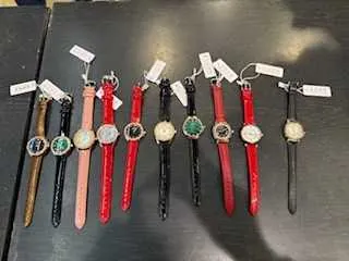 Kolekcja zegarków na rękę ułożonych w rzędzie na ciemnej powierzchni. Zegarki mają różne kolory pasków: brązowy, różowy, czerwony, czarny. Każdy zegarek posiada białą etykietę z niewidocznym tekstem. Tarcze zegarków różnią się wzorami i kolorami, w tym zielonym, białym i czarnym.