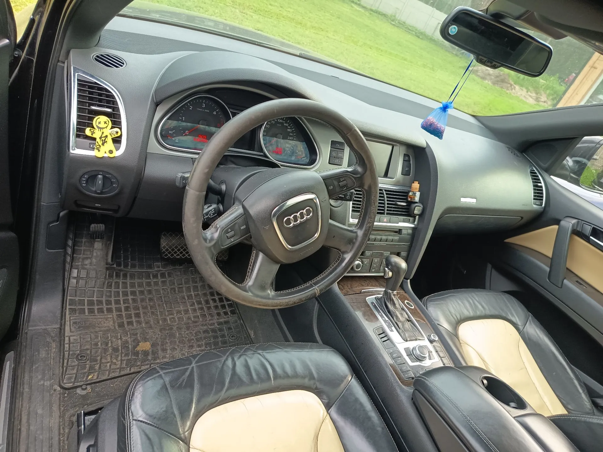 Wnętrze samochodu marki Audi widziane z perspektywy kierowcy. Widoczna jest czarna kierownica z logo czterech pierścieni, deska rozdzielcza z zegarami, lewarek zmiany biegów oraz dwa fotele wykonane z czarnej i beżowej skóry. Na lewym nawiewie przyklejone są żółte naklejki w kształcie uśmiechniętych buziek, a do lusterka wstecznego przyczepiony jest niebieski odświeżacz powietrza.