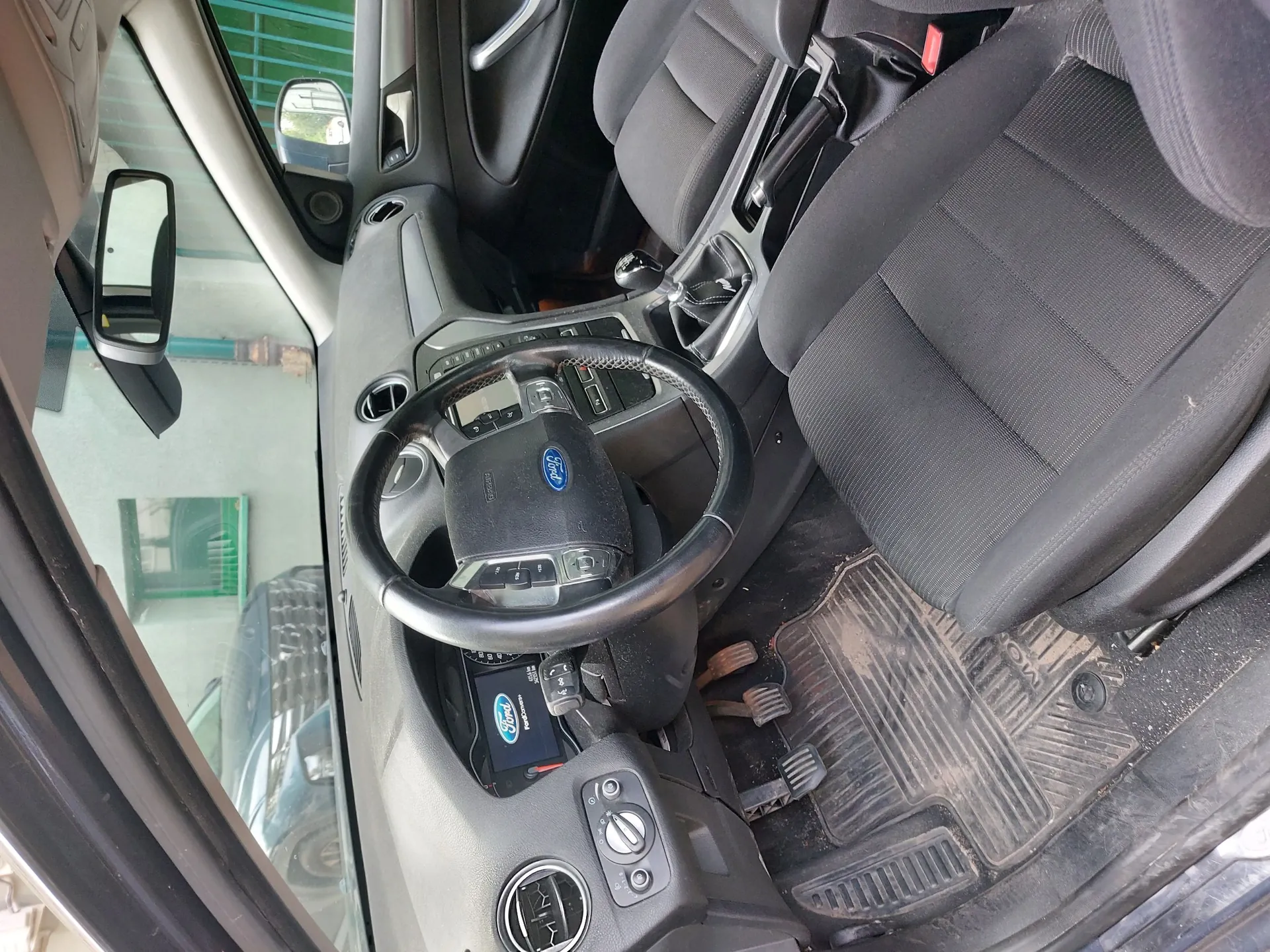 Wnętrze samochodu marki Ford widziane z perspektywy otwartych drzwi kierowcy. Widoczna jest czarna kierownica z logo Forda, deska rozdzielcza z systemem audio, lewarek zmiany biegów oraz dwa przednie fotele z ciemnoszarej tkaniny. Na podłodze znajduje się gumowa mata z widocznymi zabrudzeniami. Przez szybę widoczny jest fragment innego pojazdu i zielony płot.