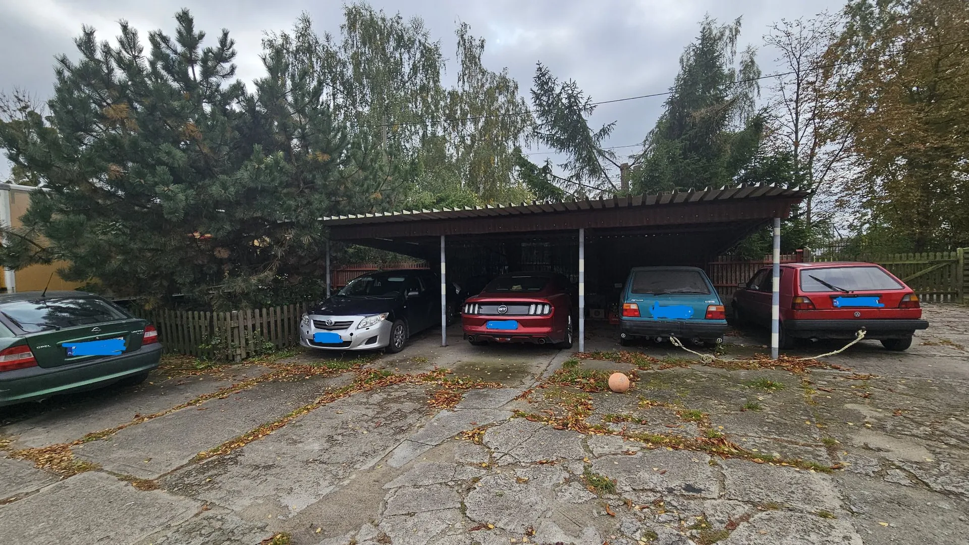 Cztery samochody zaparkowane na betonowym podjeździe z liśćmi. Trzy z nich znajdują się pod prostą wiatą z ciemnym dachem: srebrny sedan, czerwony Mustang oraz niebieski samochód. Czwarty, ciemnozielony samochód, stoi po lewej stronie poza wiatą. W tle widoczne są gęste, zielone drzewa i drewniany płot pod zachmurzonym niebem.
