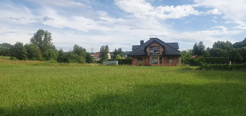 Dom w trakcie budowy z czerwonej cegły z ciemnym dachem, położony na dużym zielonym polu. W tle drzewa, inne budynki oraz niebo z białymi chmurami. Horyzont jest poziomy, a całość ustawiona pionowo.