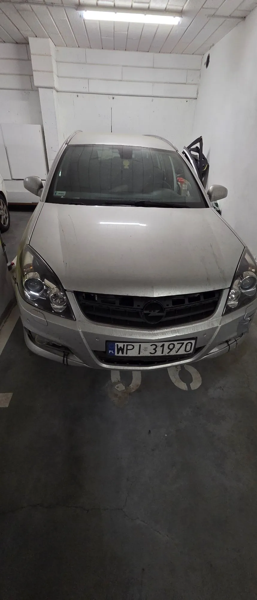 Srebrny samochód marki Opel zaparkowany w białym garażu. Widoczny jest przód pojazdu z polską tablicą rejestracyjną o numerze WPI 31970. Samochód ma widoczne uszkodzenia z przodu, w tym pęknięty zderzak po prawej stronie (patrząc od przodu).
