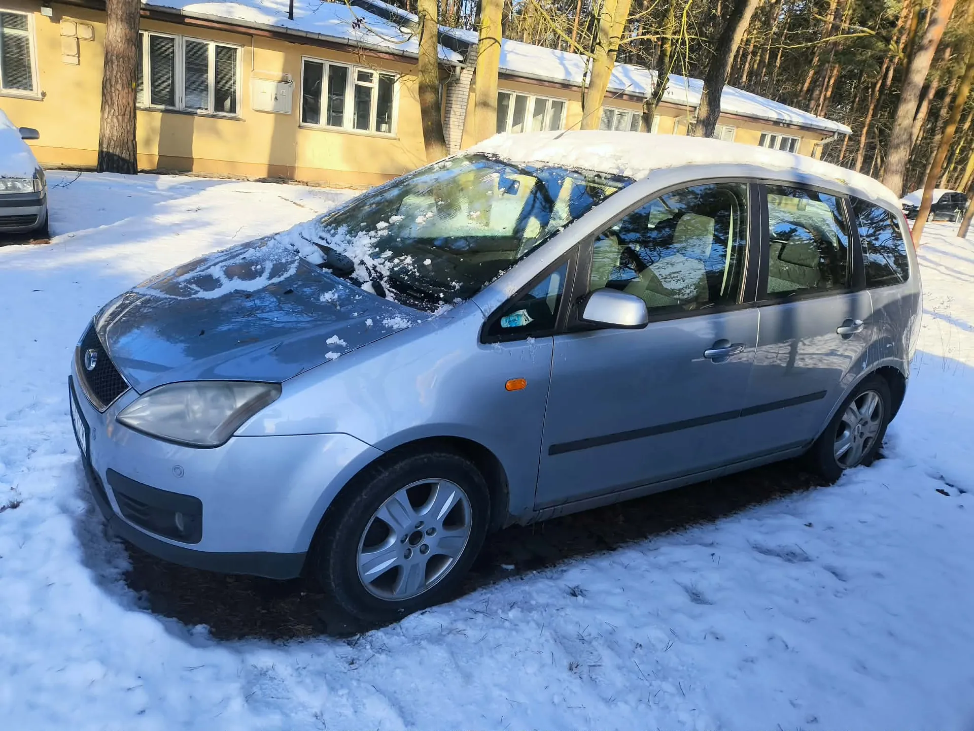 Srebrny samochód osobowy zaparkowany na śnieżnej powierzchni. Na dachu i masku pojazdu widoczny jest śnieg. W tle żółty budynek z oknami i śniegiem na dachu, otoczony drzewami bez liści. Po lewej stronie częściowo widoczny inny samochód. Teren pokryty śniegiem, słońce oświetla scenę, rzucając cienie.