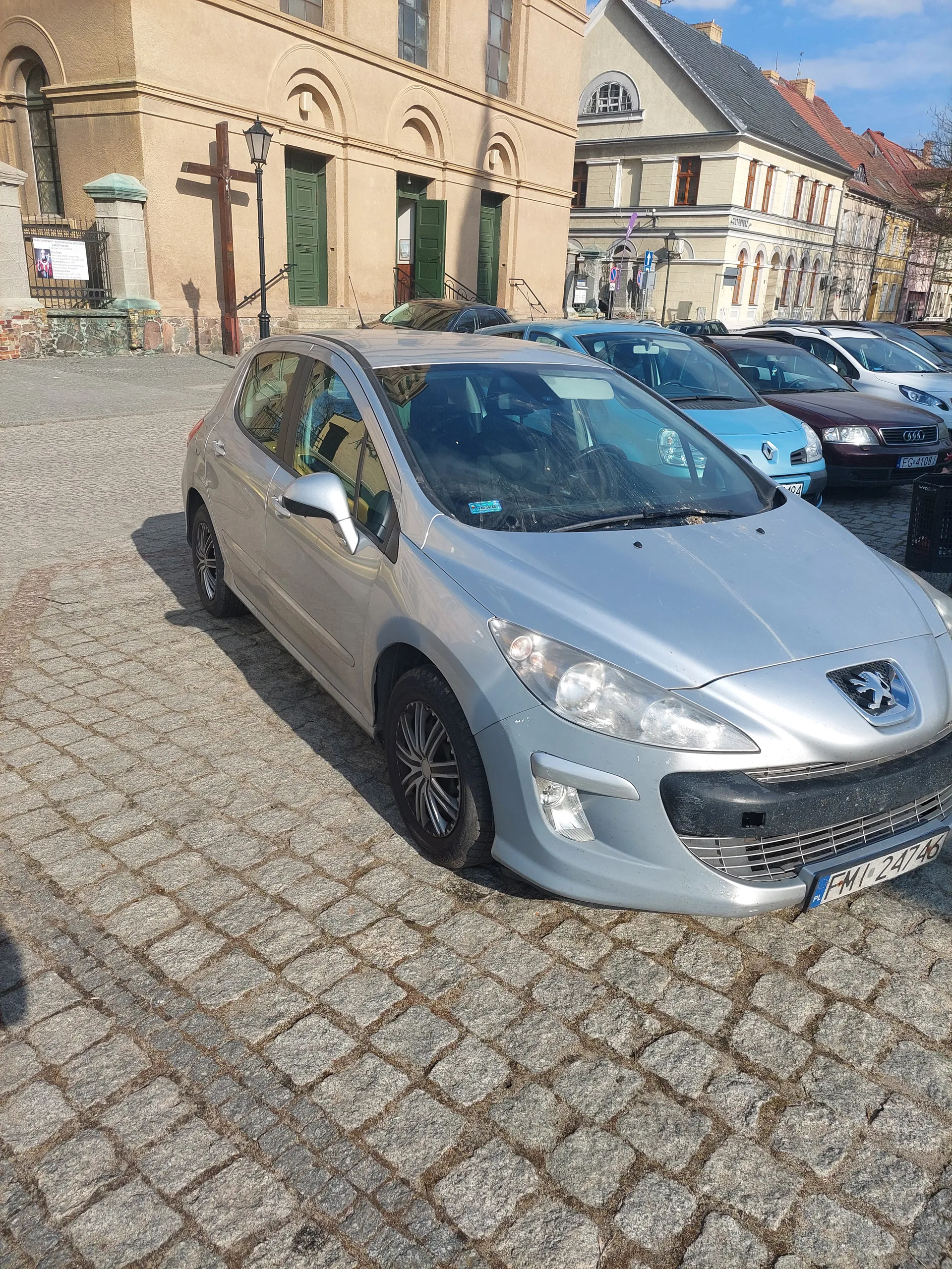 Srebrny samochód marki Peugeot zaparkowany na brukowanej ulicy. Widoczna tablica rejestracyjna FWL 2476. W tle inne zaparkowane samochody, budynki z architekturą europejską, w tym budynek z zielonymi drzwiami i krzyżem. Jasne niebo z nielicznymi chmurami.