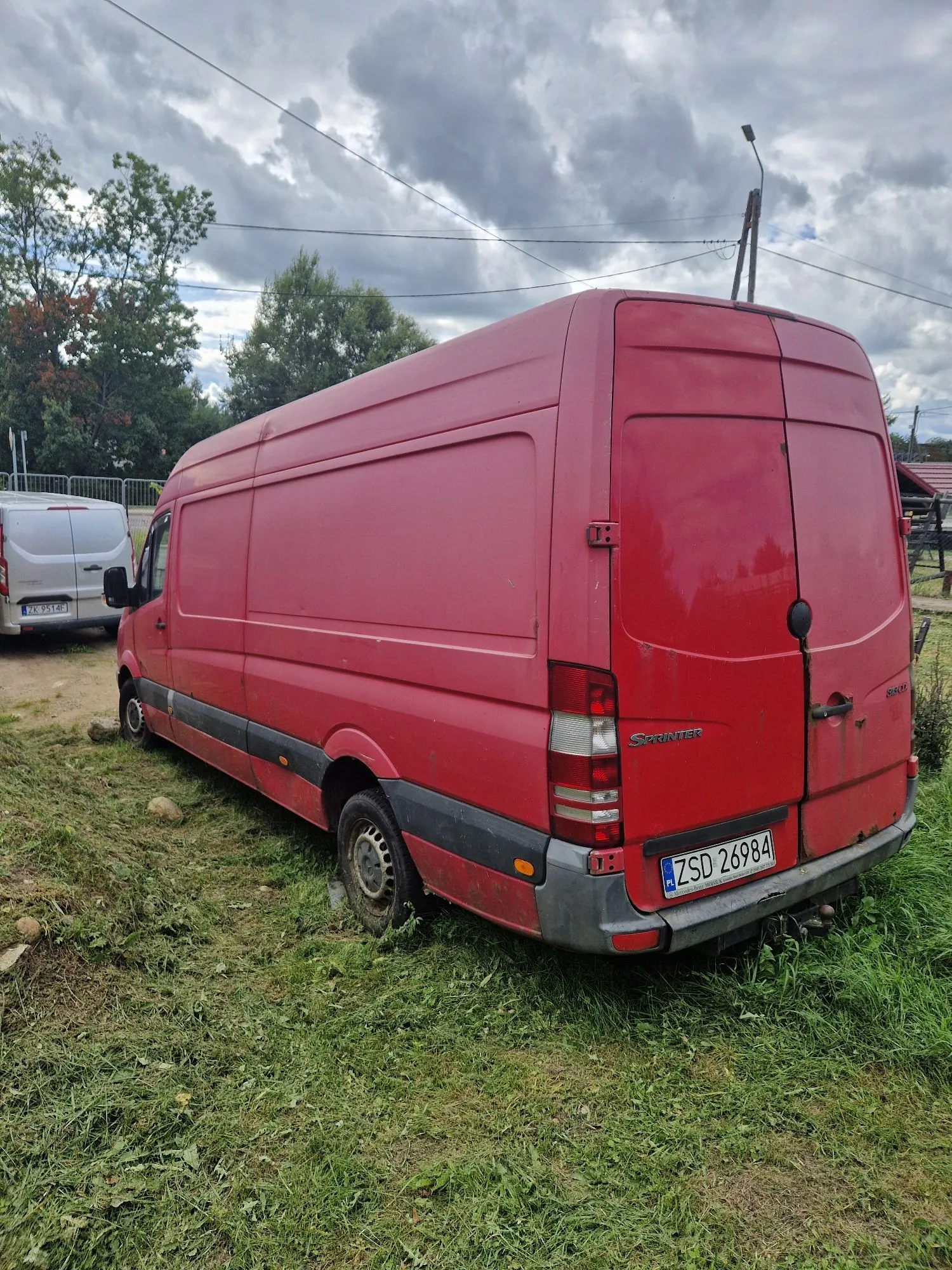 Czerwony furgon marki Mercedes-Benz Sprinter zaparkowany na trawiastym terenie. Pojazd jest widoczny z tyłu i z boku. Na tylnych drzwiach znajduje się napis 'SPRINTER' oraz polska tablica rejestracyjna o numerze 'ZSD 26984'. W tle widoczny jest inny, biały samochód dostawczy, drzewa oraz zachmurzone niebo.
