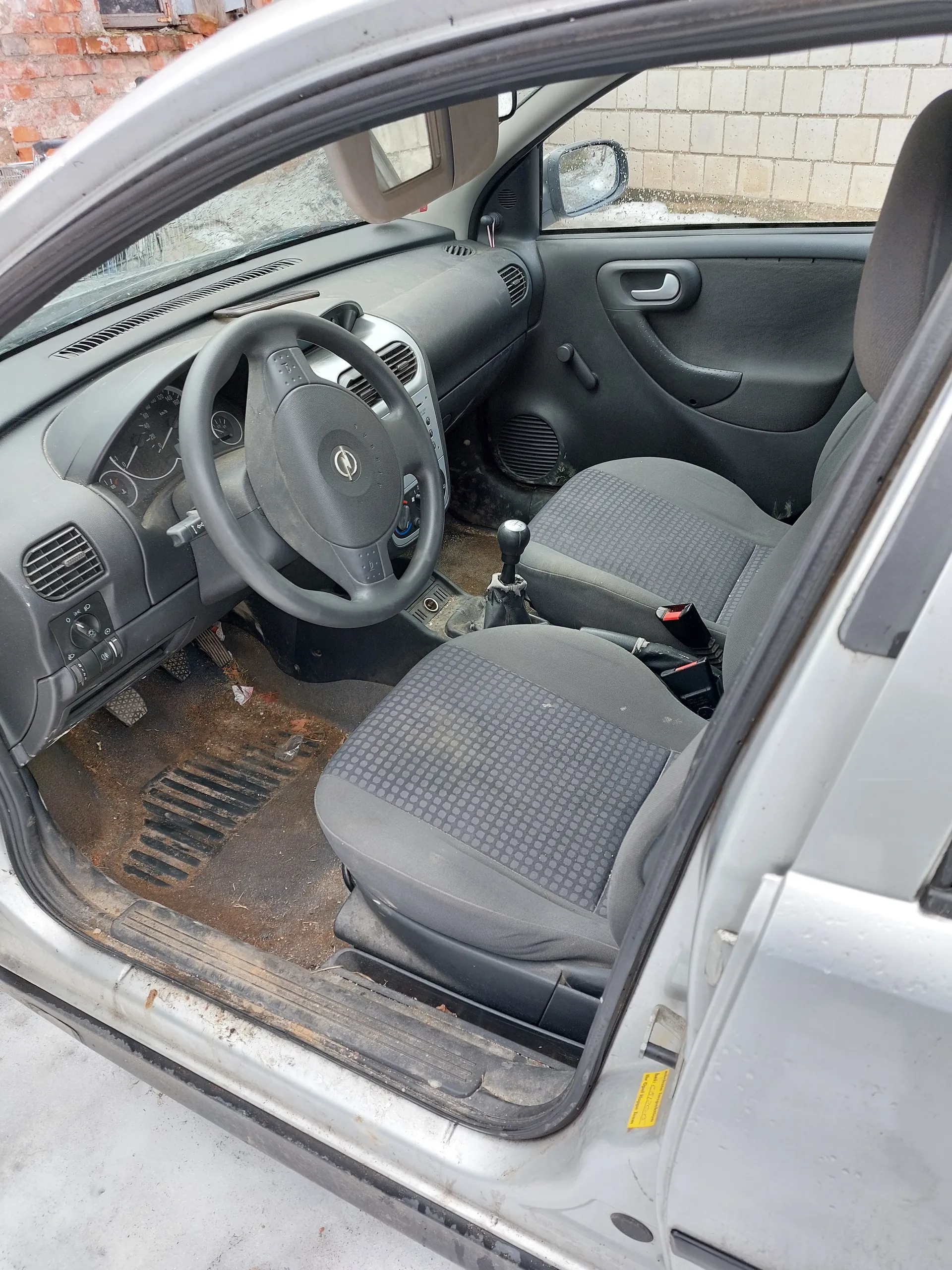 Wnętrze samochodu z otwartą drzwiami od strony kierowcy. Widoczne są kierownica z logo, deska rozdzielcza z wskaźnikami, dwa przednie siedzenia w szaro-czarnym wzorze, brudna podłoga z resztkami śmieci i zniszczonym dywanikiem. W tle za przednią szybą widać ceglaną i betonową ścianę. Drzwi samochodu są srebrne, a na ich krawędzi znajduje się żółty naklejony znaczek.